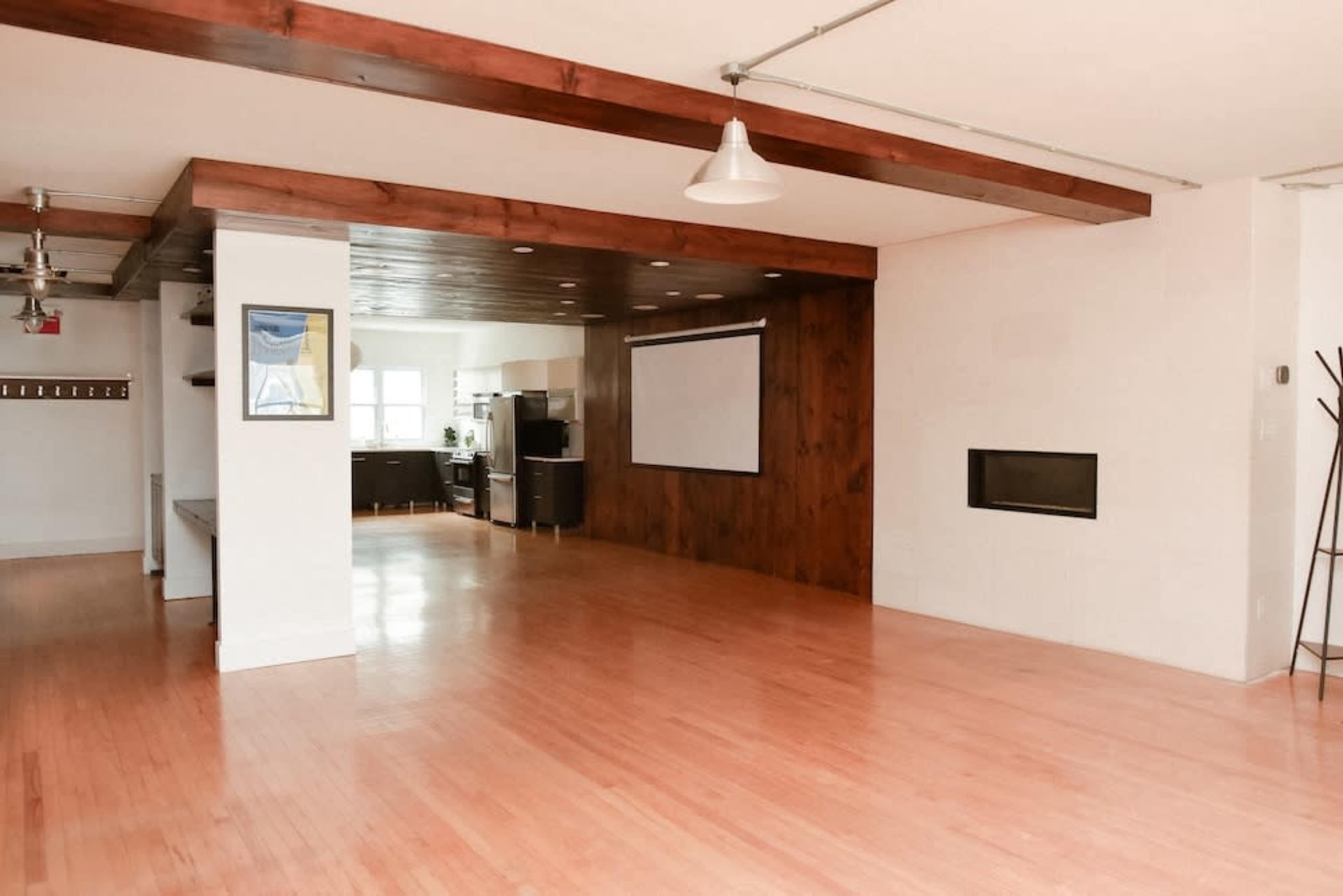 The image shows a spacious, open-concept room with wooden beams, a wall-mounted projector screen, and a modern kitchen area visible in the background.
