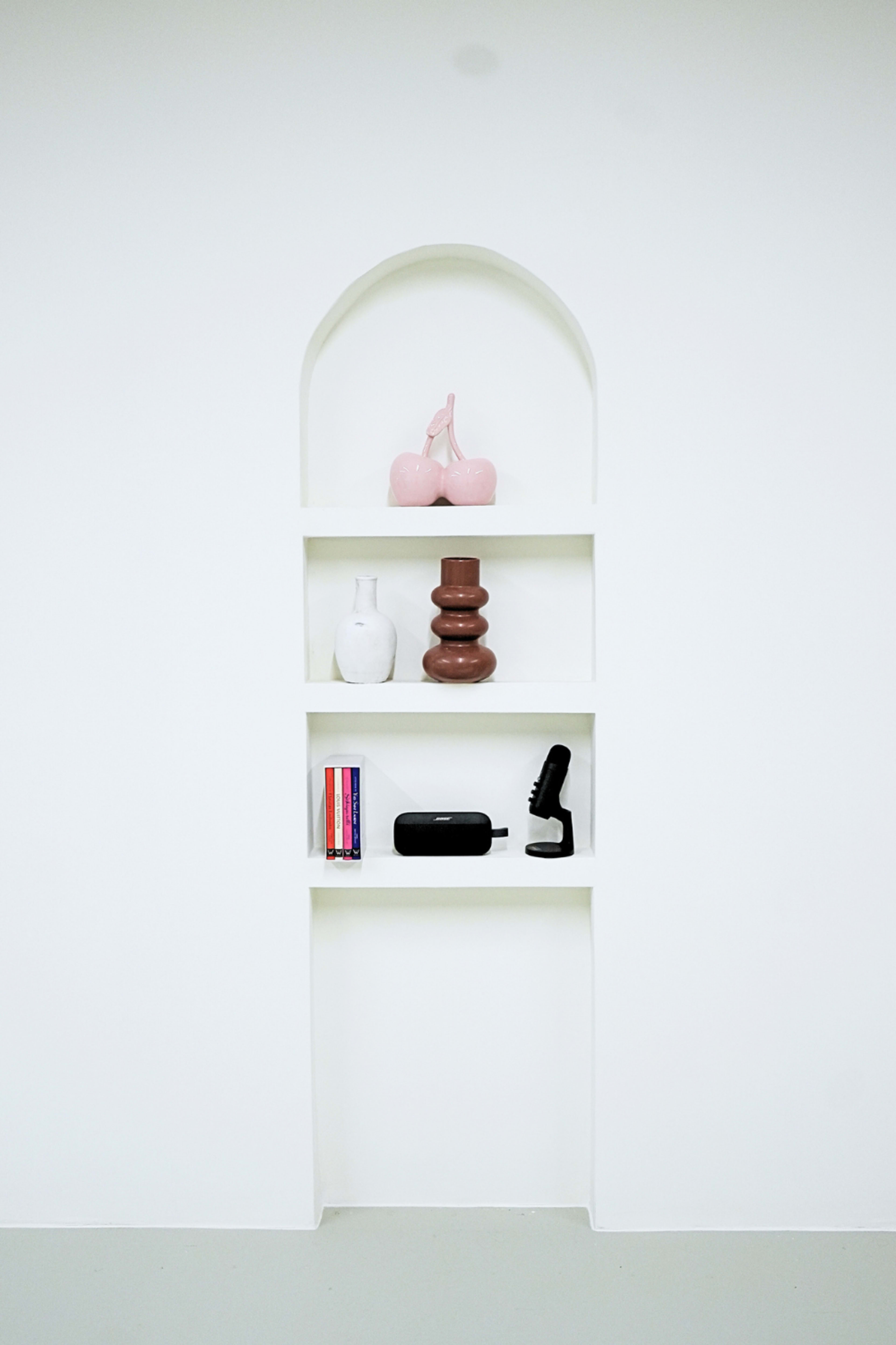 The image features a shallow wall niche with three shelves displaying various decorative objects, including a pink vase, a brown sculpture, a white vase, a stack of books, and a black speaker.