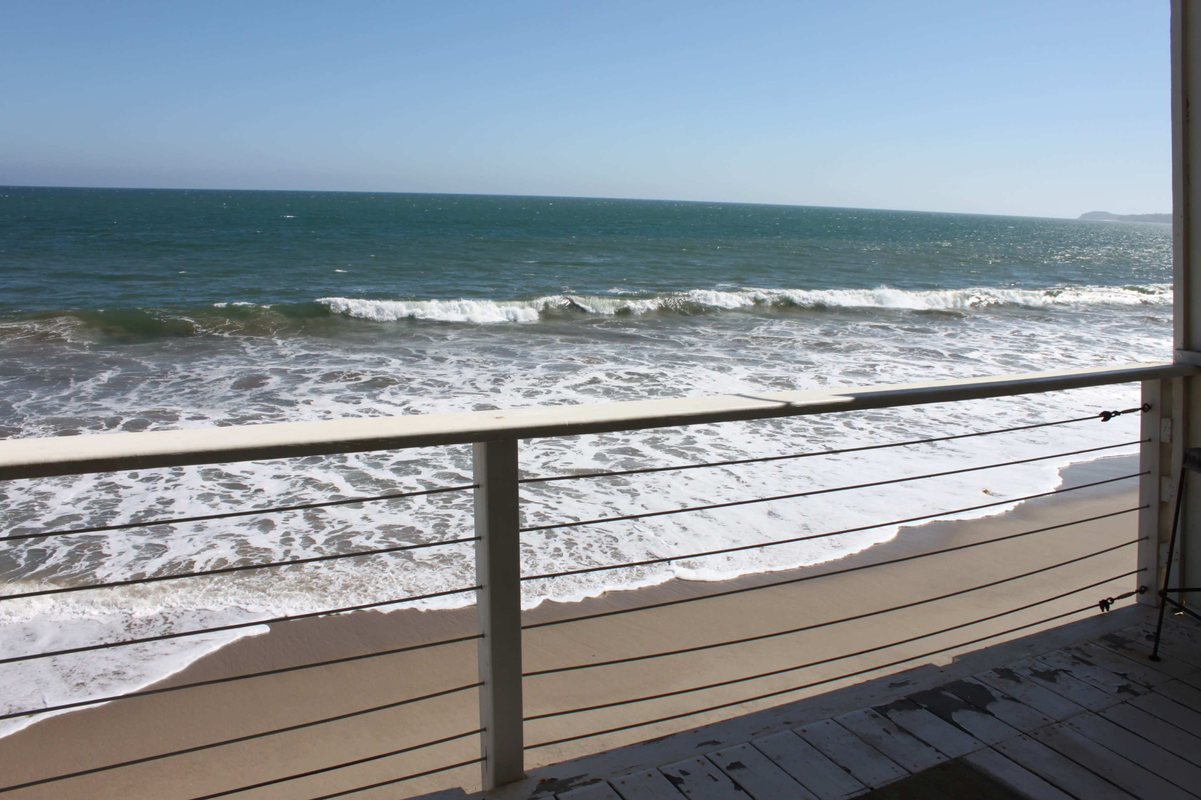 The image shows a view of waves crashing against a sandy beach, with a balcony railing in the foreground.