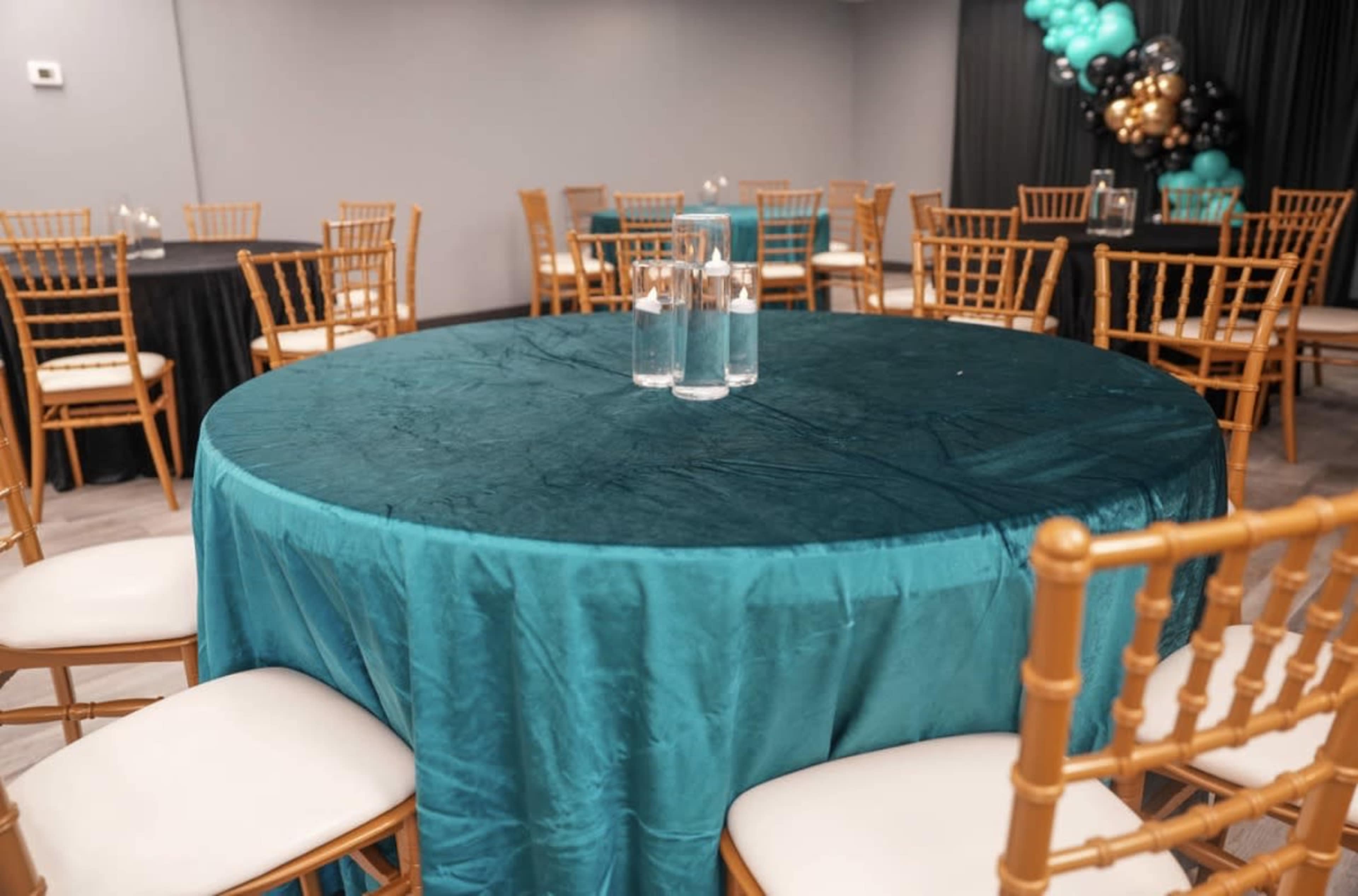 A round table with a teal tablecloth is set in a banquet hall, surrounded by wooden chairs and decorated with clear glasses of water on the table.