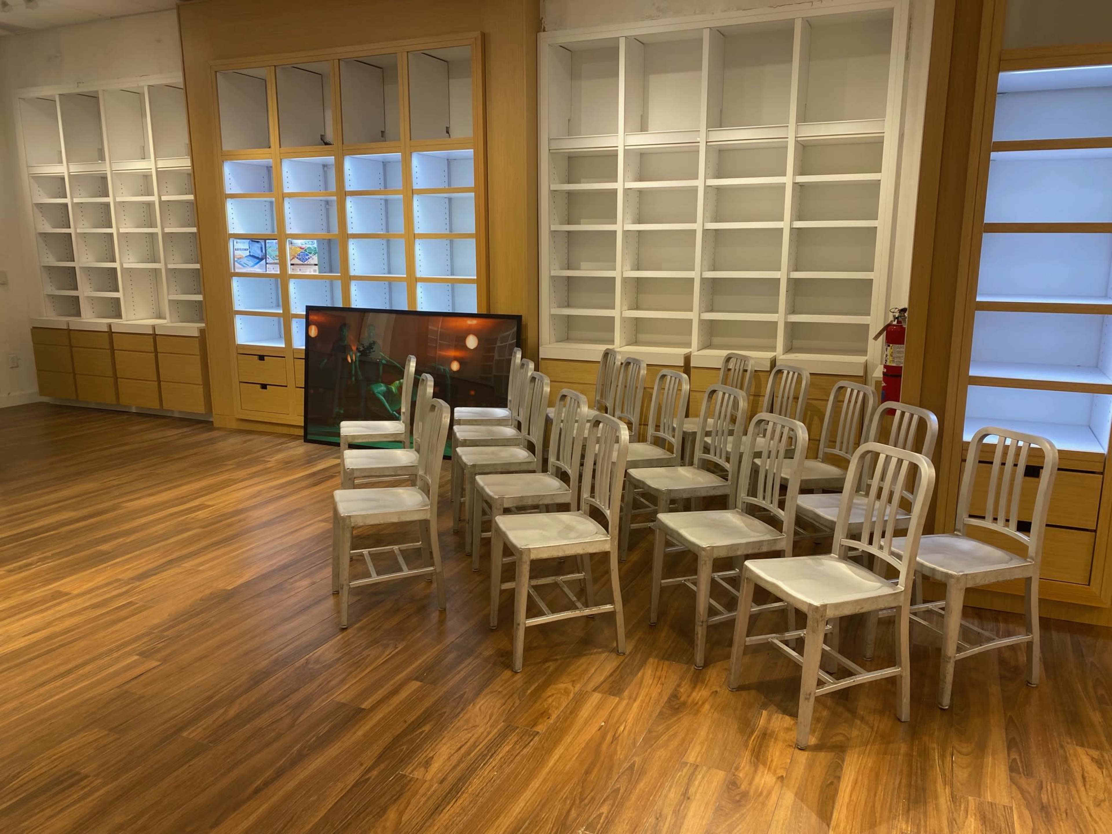A series of metal chairs arranged in two rows in a room with wooden flooring and empty shelves.