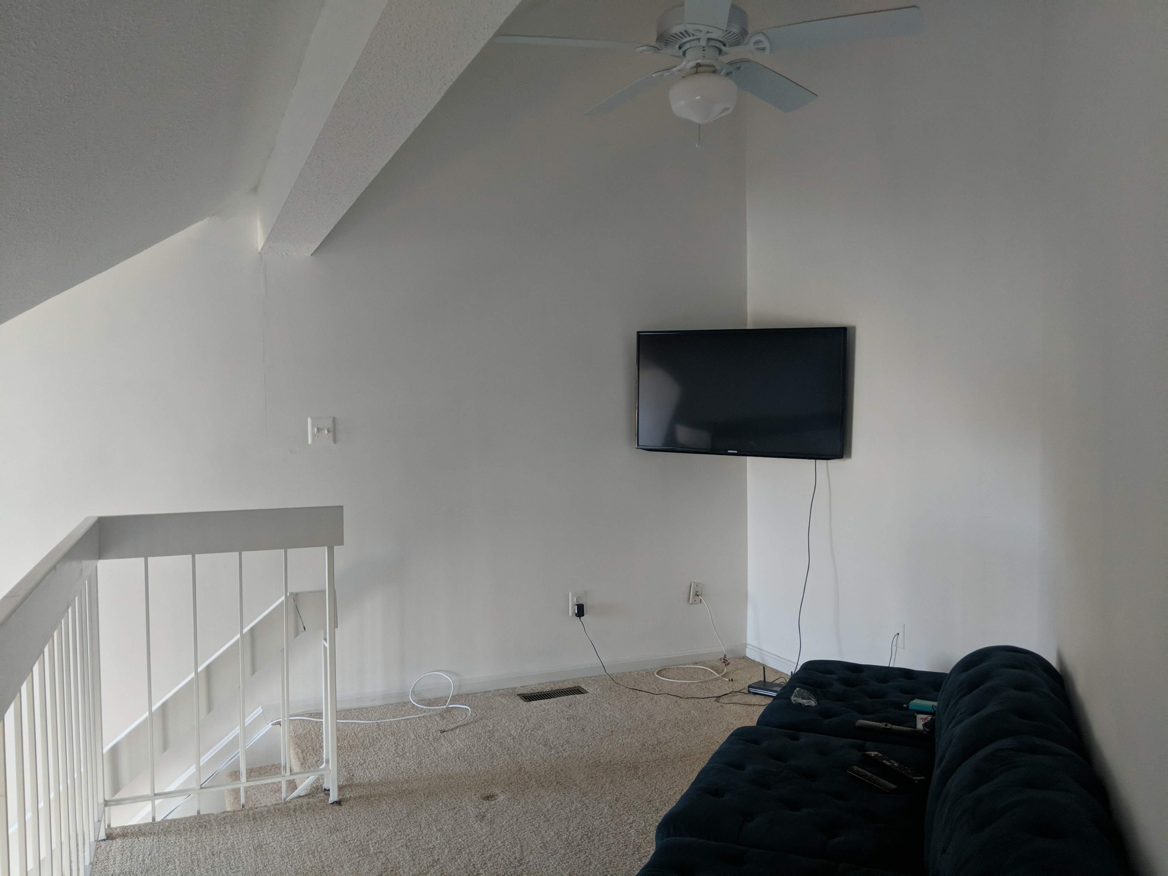 A sparsely furnished living room with a dark couch, a mounted television on the wall, and a staircase leading to another level.