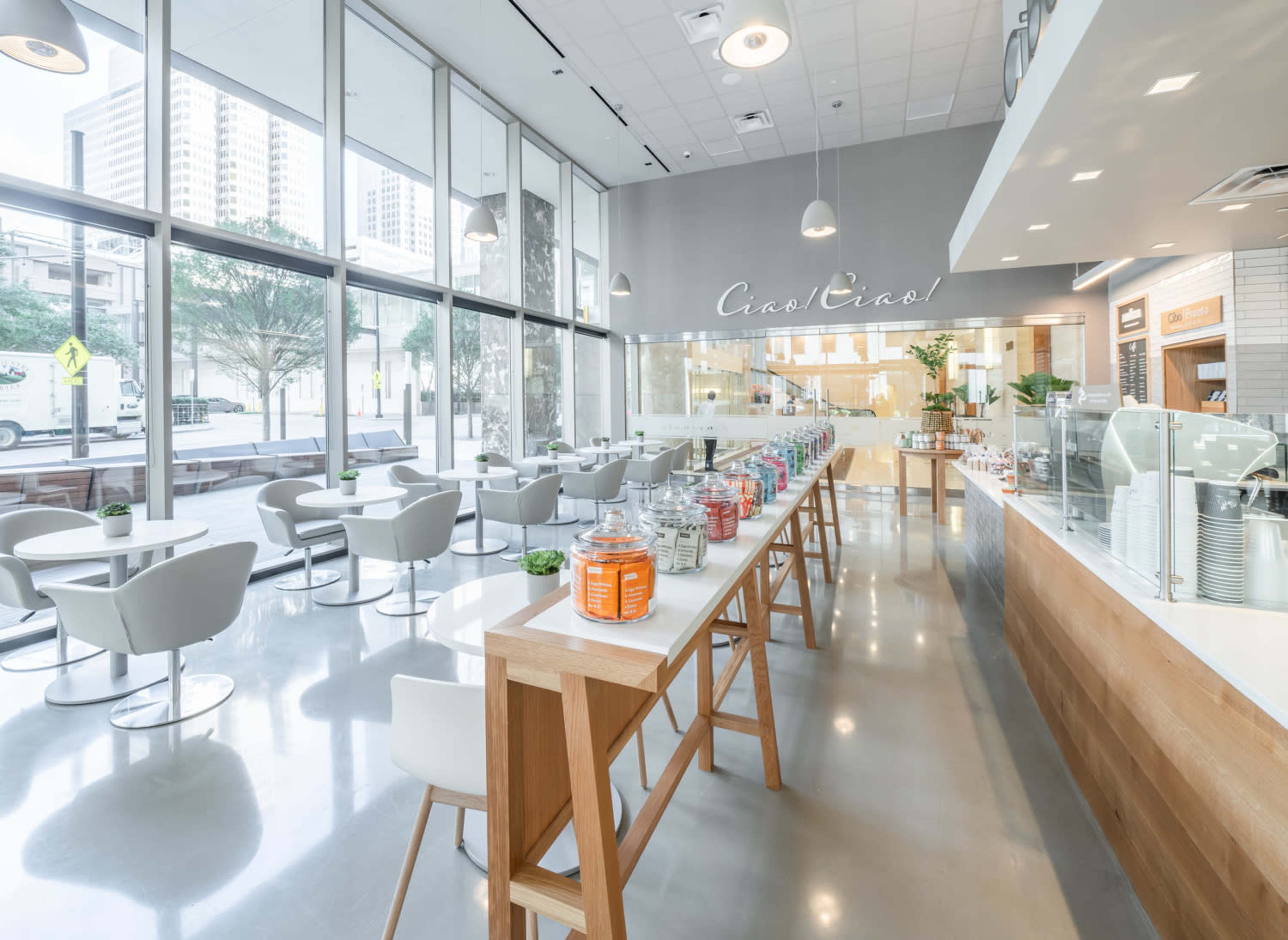 The image shows a modern café with large windows, featuring seating areas and a display of jars on a counter.