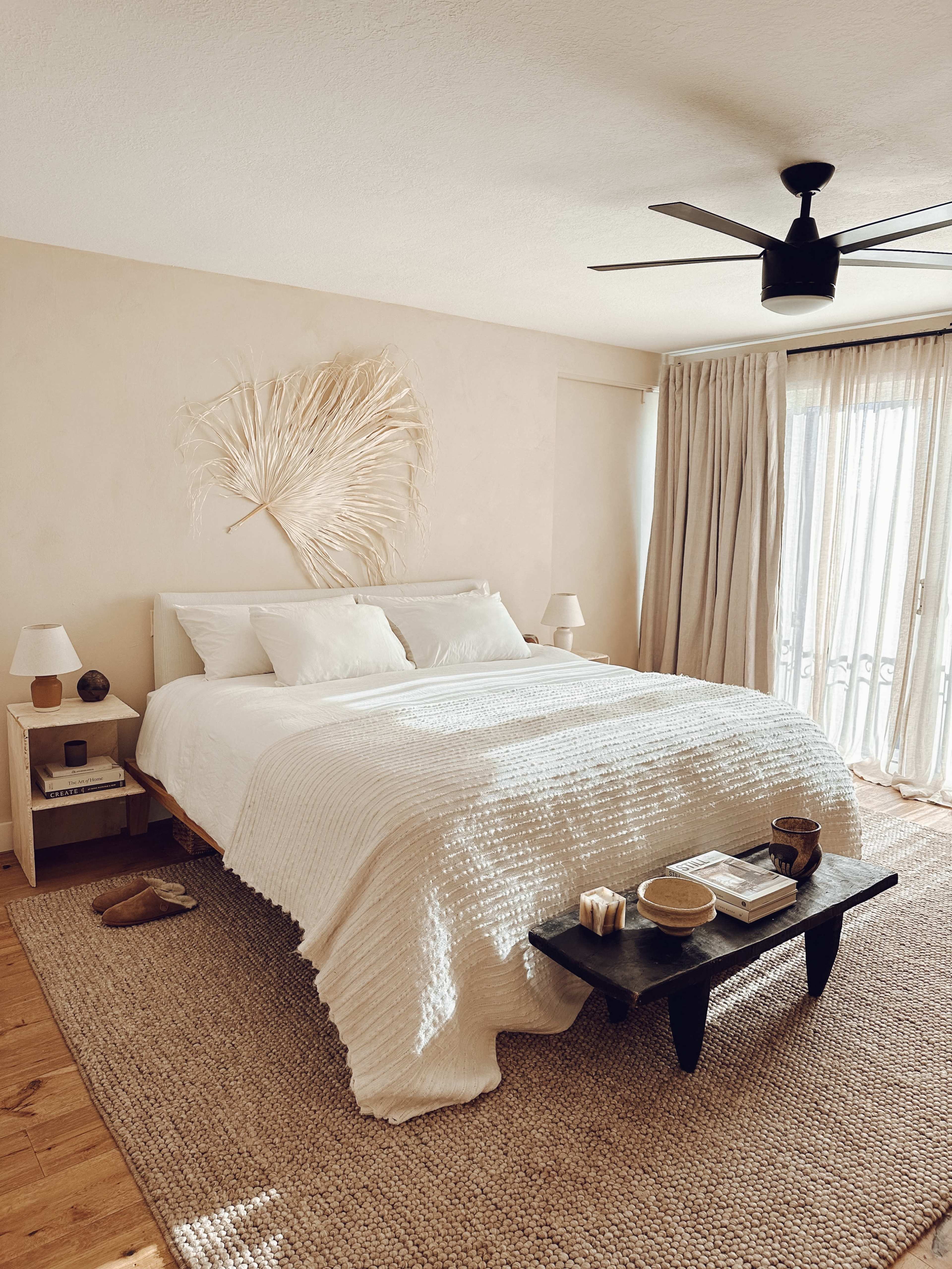 A light-toned bedroom featuring a bed with white bedding, a small black coffee table, and large sliding glass doors covered with sheer curtains.