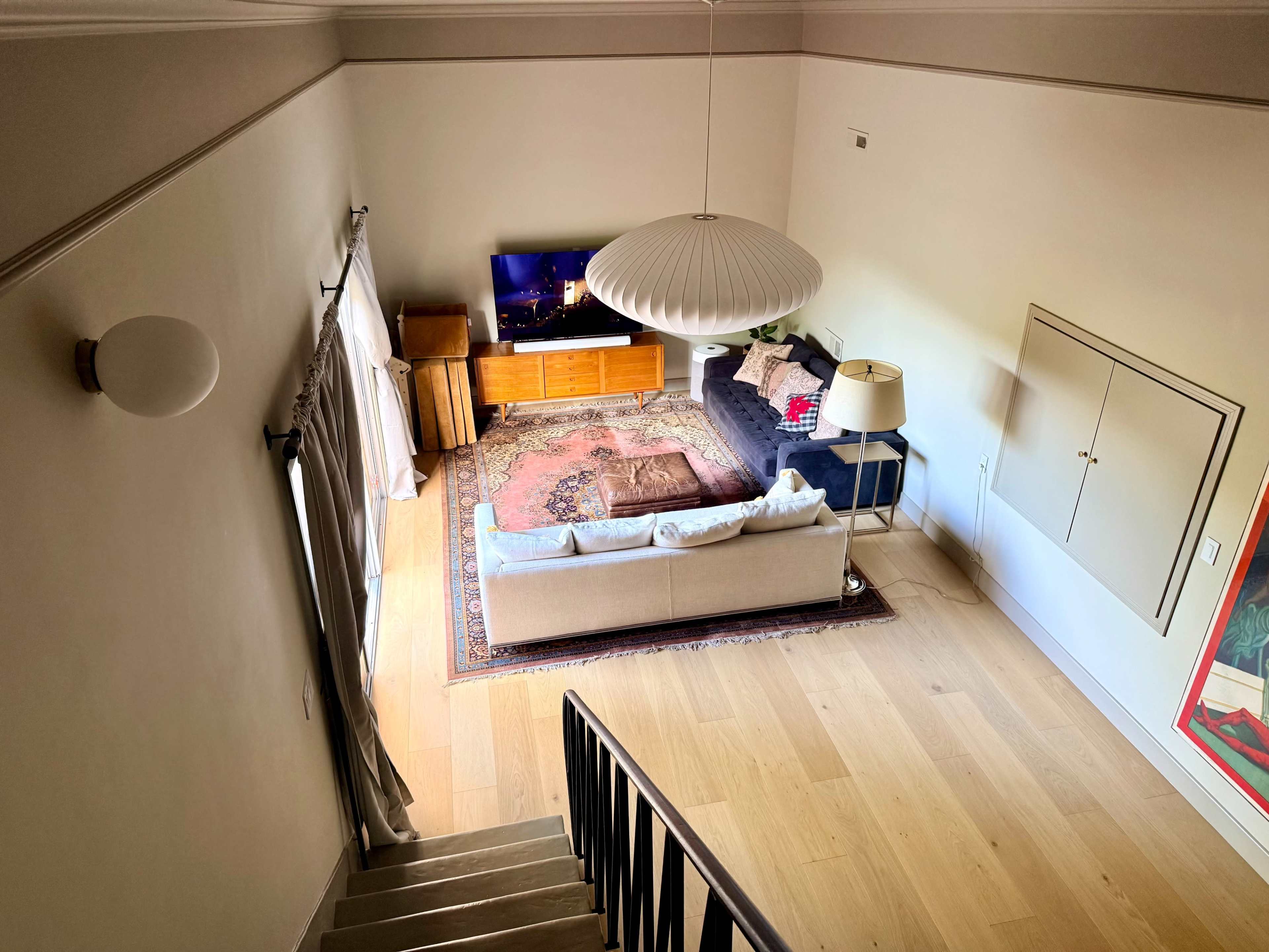The image shows a living room viewed from an upper level, featuring a couch, a television, a rug, a lamp, and a wooden cabinet against the wall.