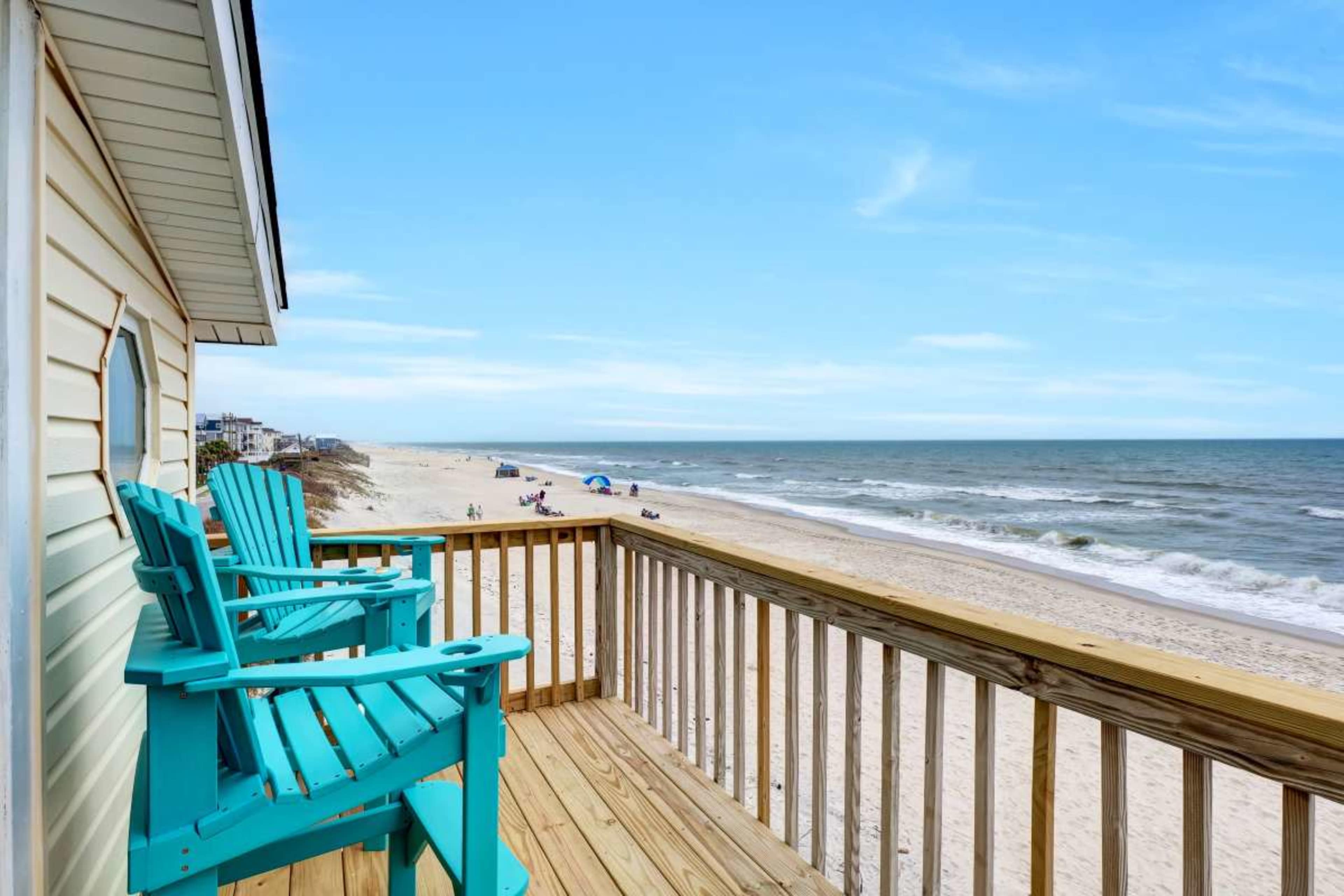 A wooden deck with turquoise chairs overlooks a sandy beach and the ocean in the distance.