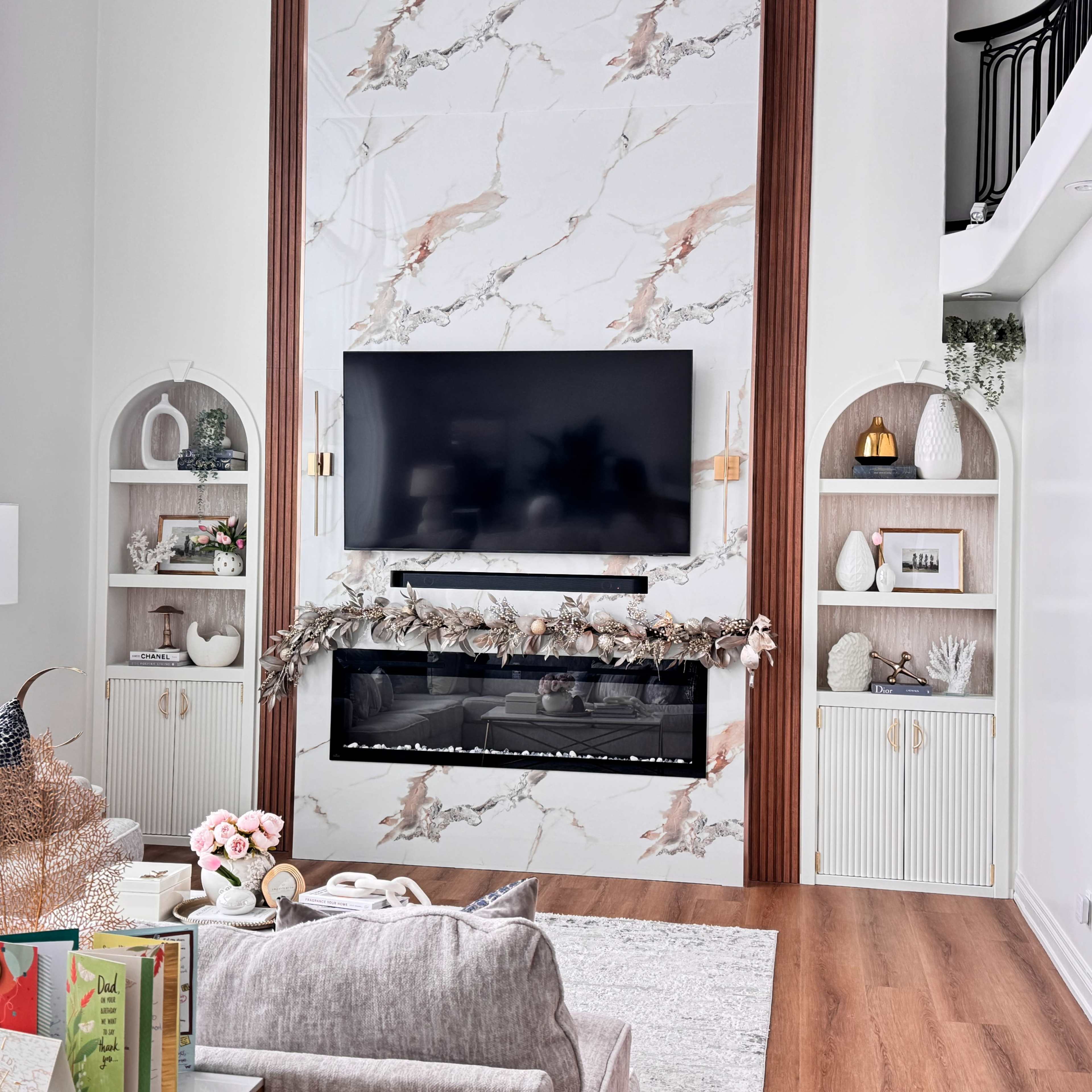 The image features a modern living room with a large wall-mounted television above a sleek electric fireplace, flanked by built-in shelves displaying decorative items and books.