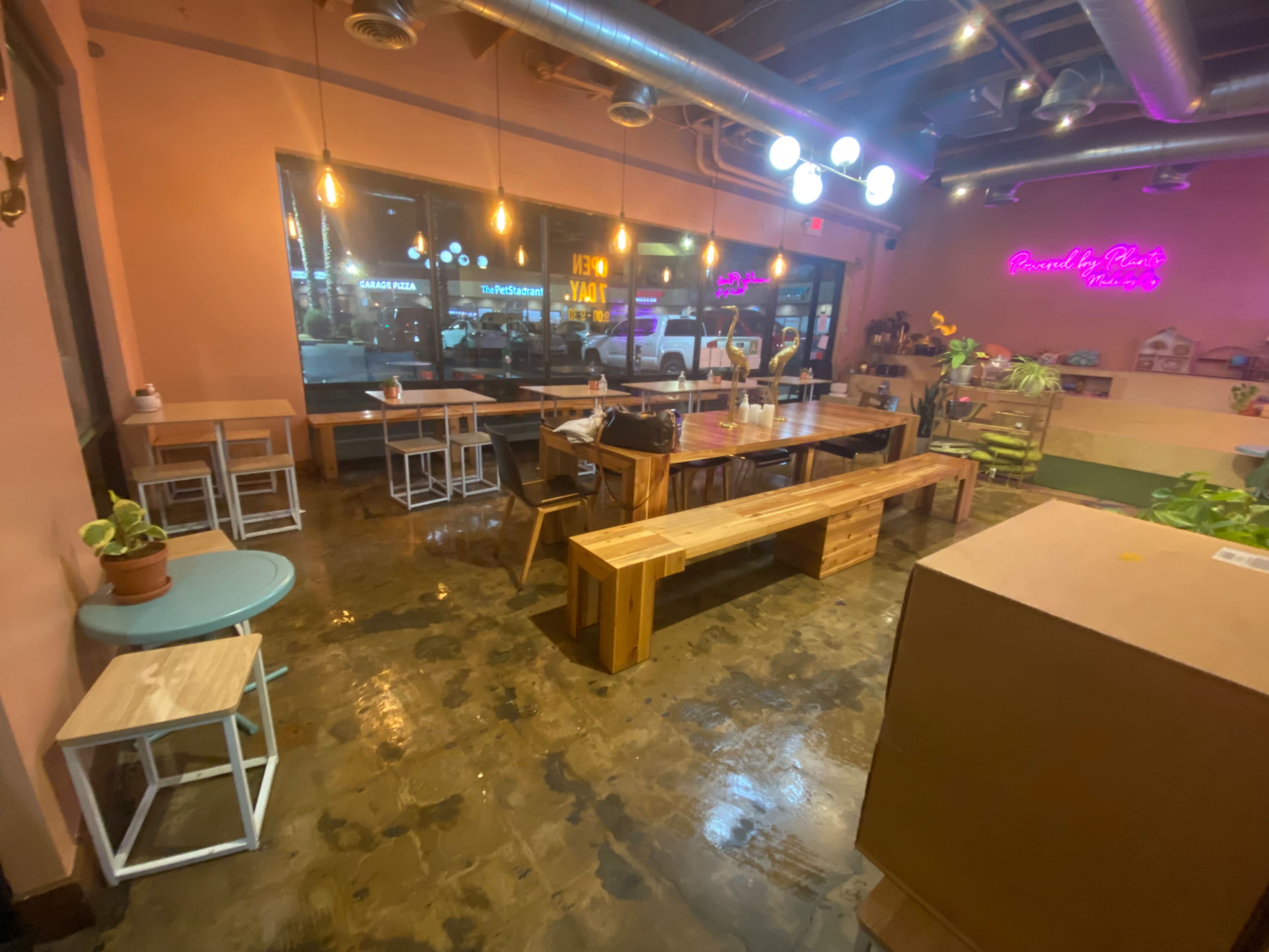 A contemporary café interior with wooden tables and benches, dim lighting, and large windows displaying neon signs outside.