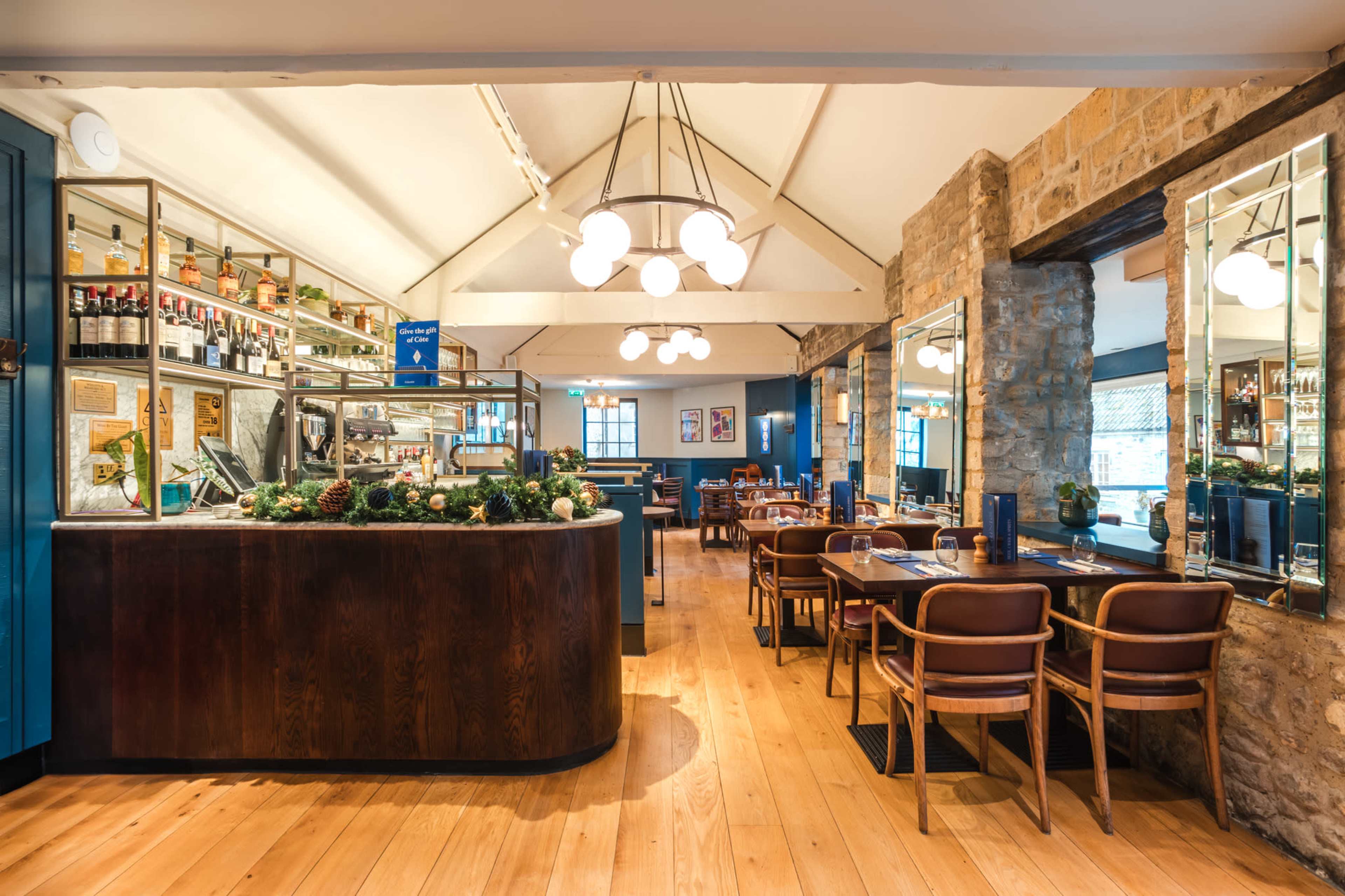 The image shows a modern café interior with a wooden counter, tables, and large windows, featuring warm lighting and stone walls.
