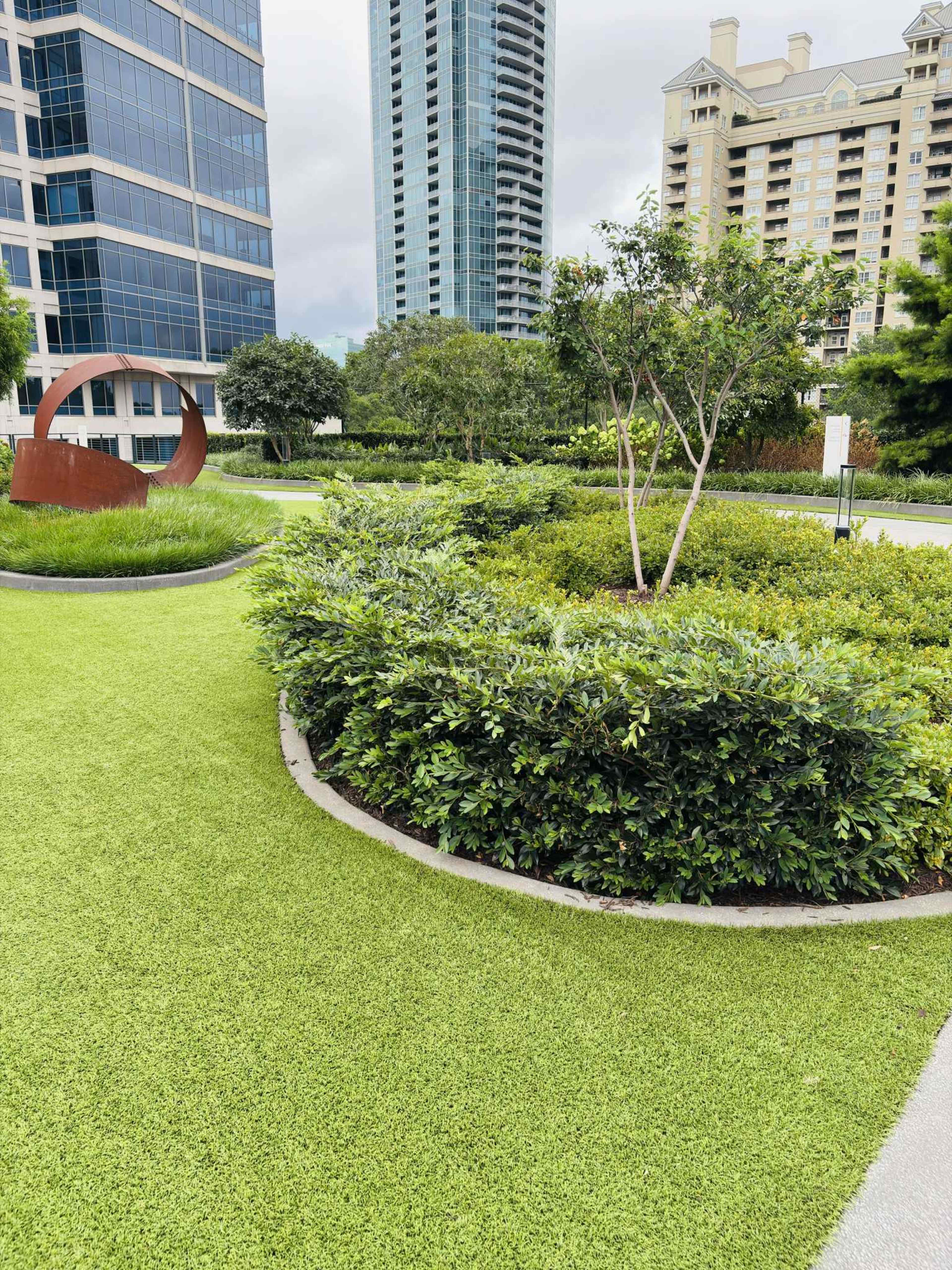 The scene features a landscaped area with green grass, circular garden beds, and modern buildings in the background.