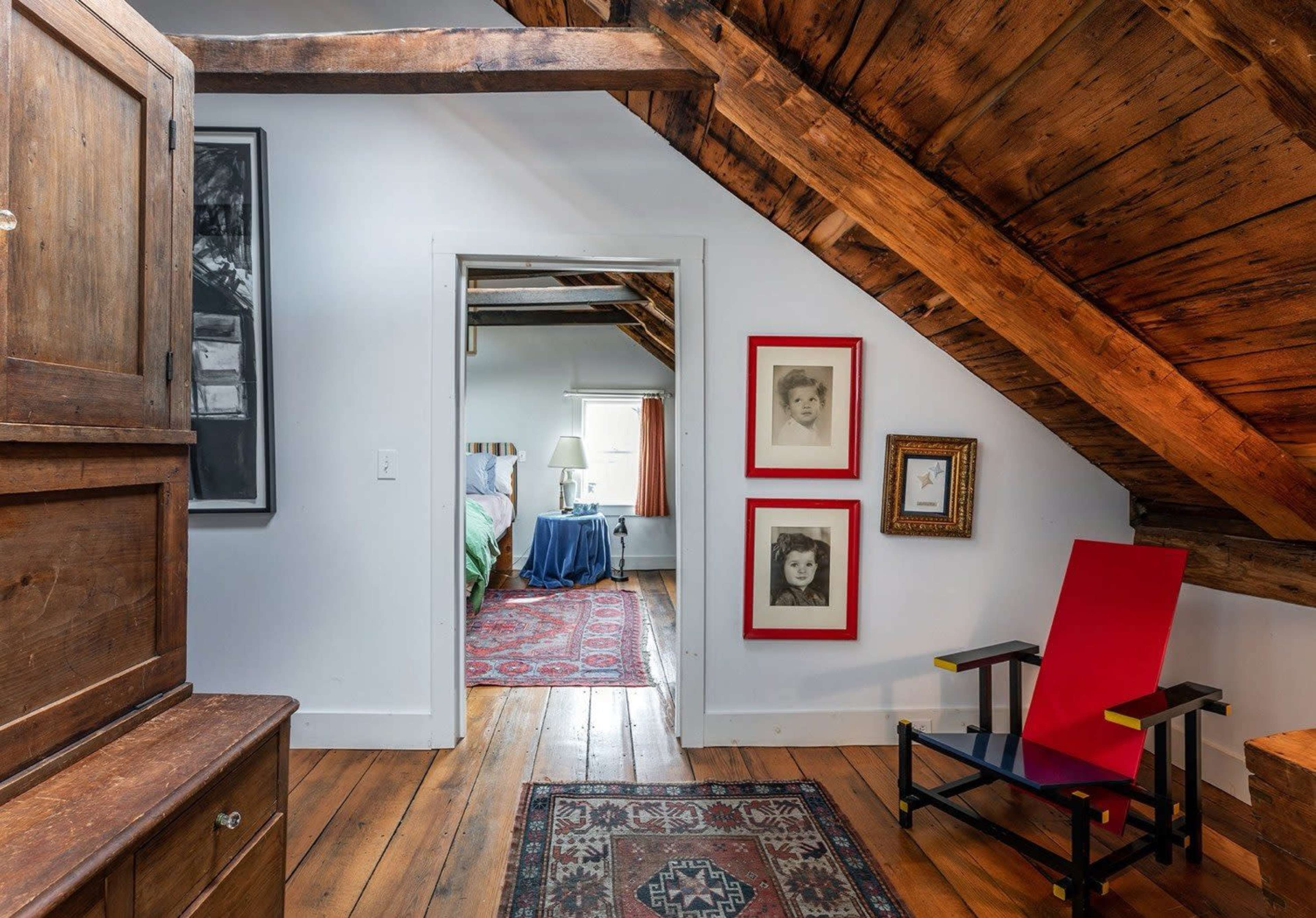 The image shows a rustic attic room with exposed wooden beams, a red chair, and framed pictures on the wall, leading to a bedroom in the background.