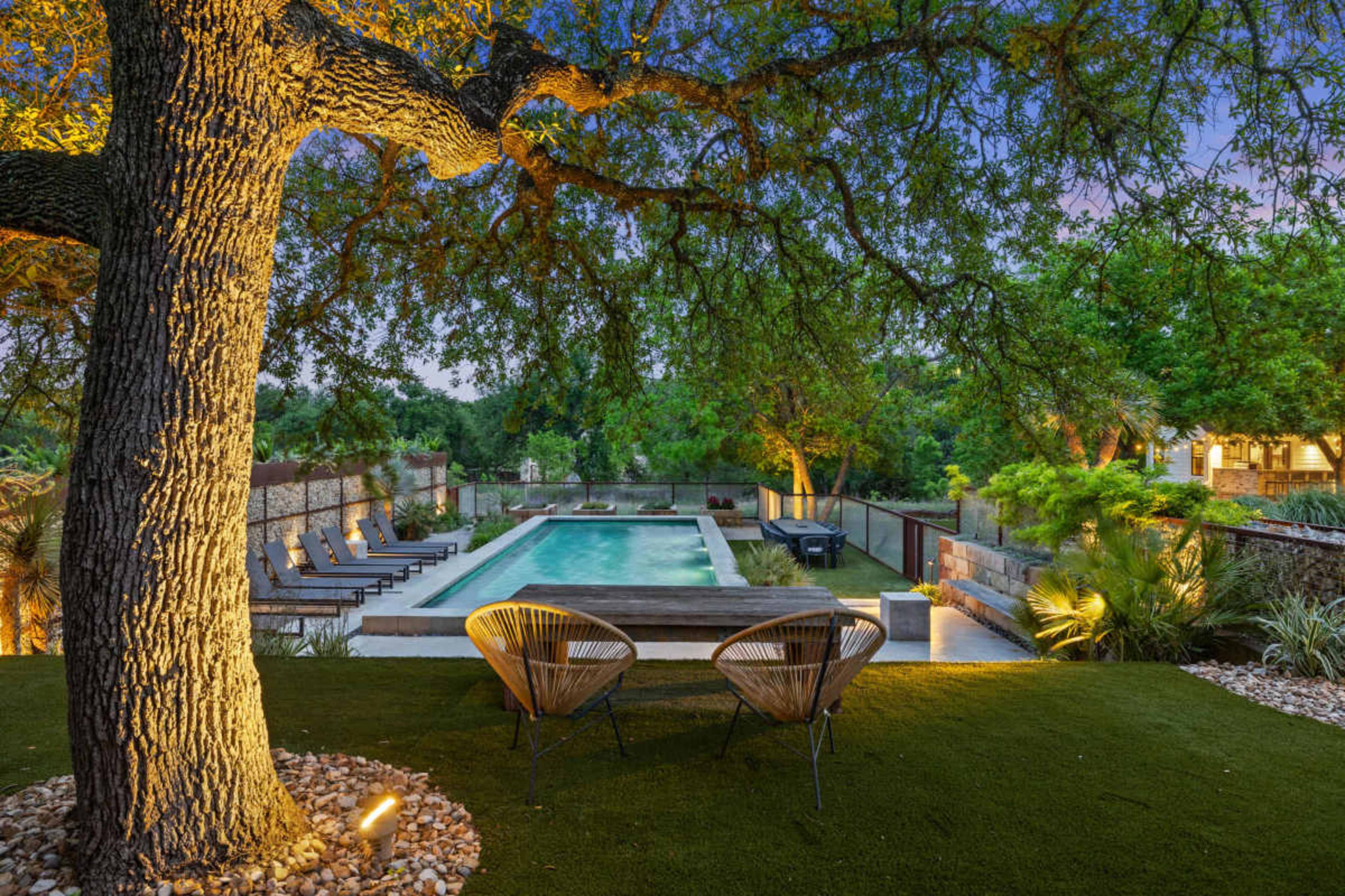 The image shows a landscaped outdoor area featuring a swimming pool surrounded by lounge chairs, a tree, and evening lighting.