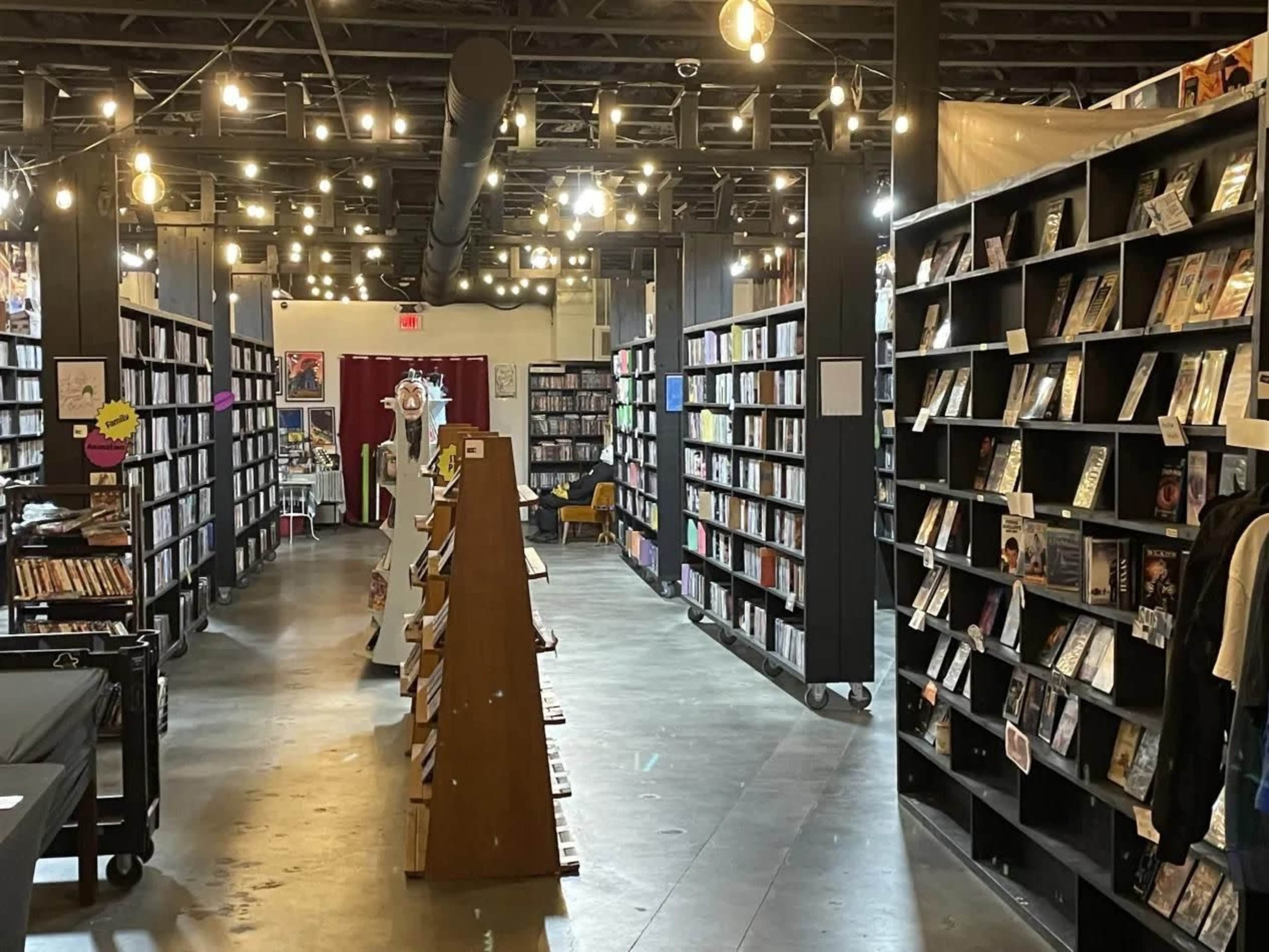 The image shows a well-lit bookstore with tall bookshelves arranged in rows, and a display of books and magazines in the center aisle.