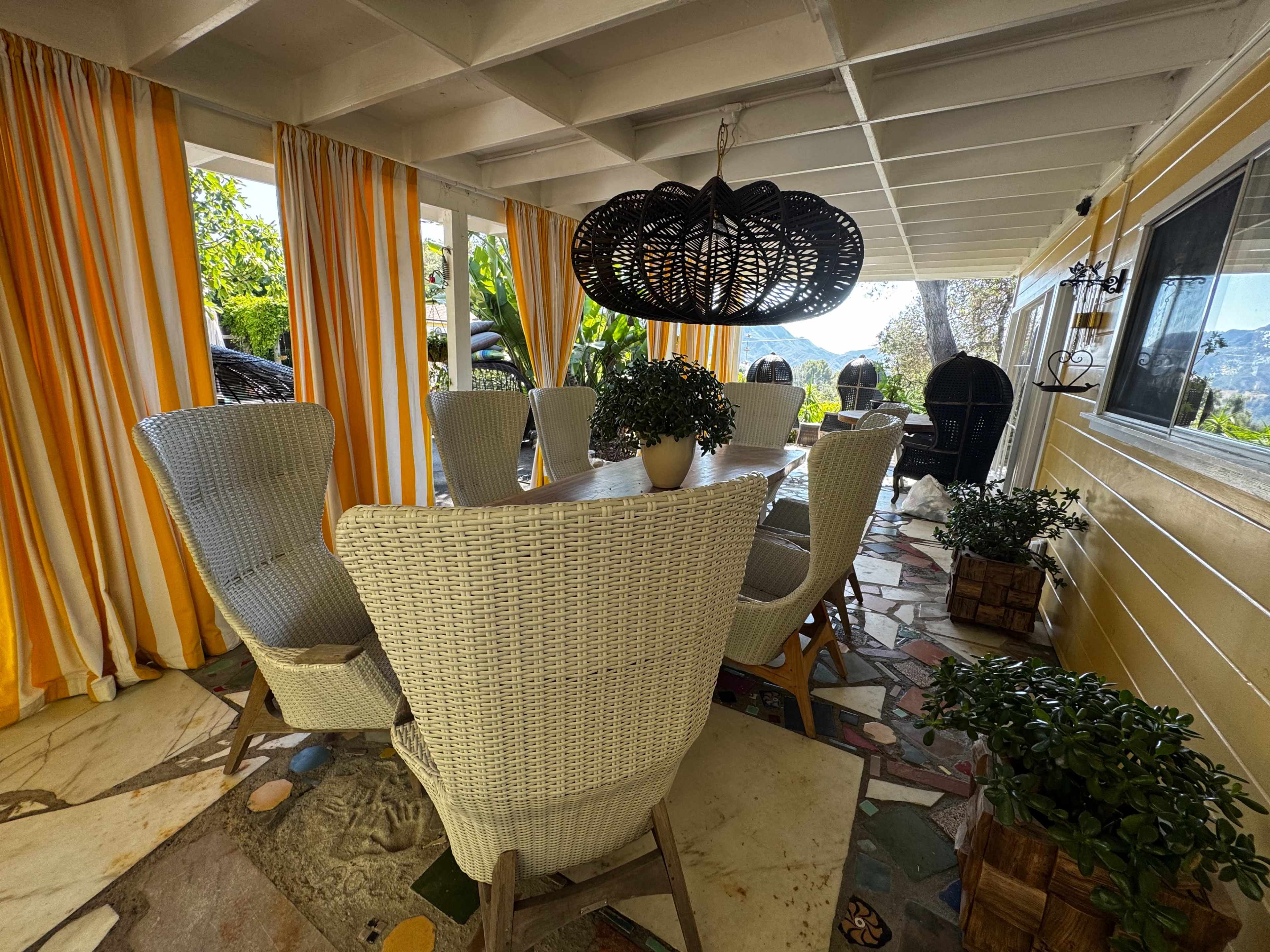 The image shows a stylish outdoor dining area with a large table surrounded by white wicker chairs, adorned with orange and yellow striped curtains and lush greenery.