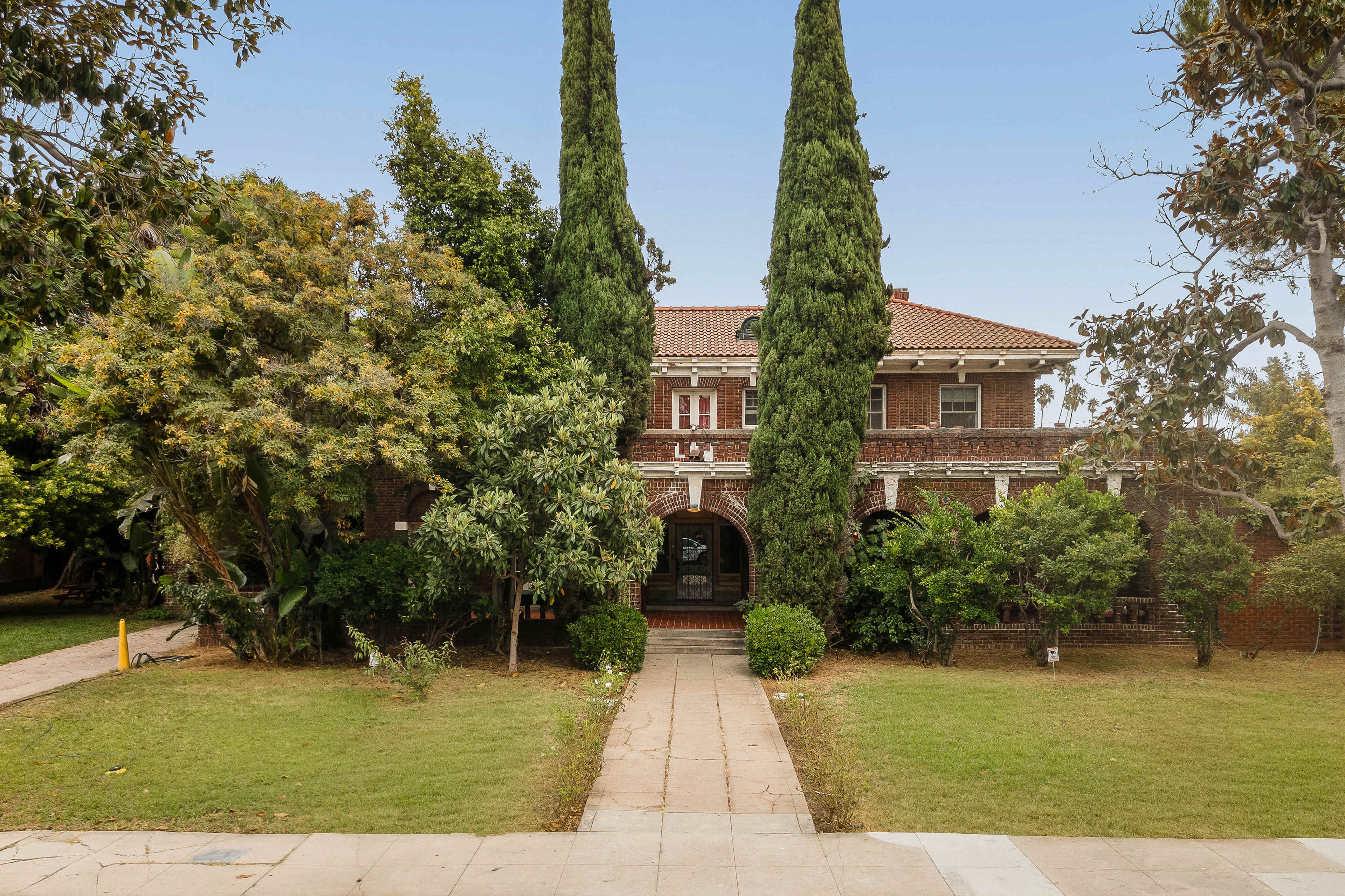 The image shows a large brick building with a red tiled roof, flanked by tall, narrow trees and surrounded by lush greenery.