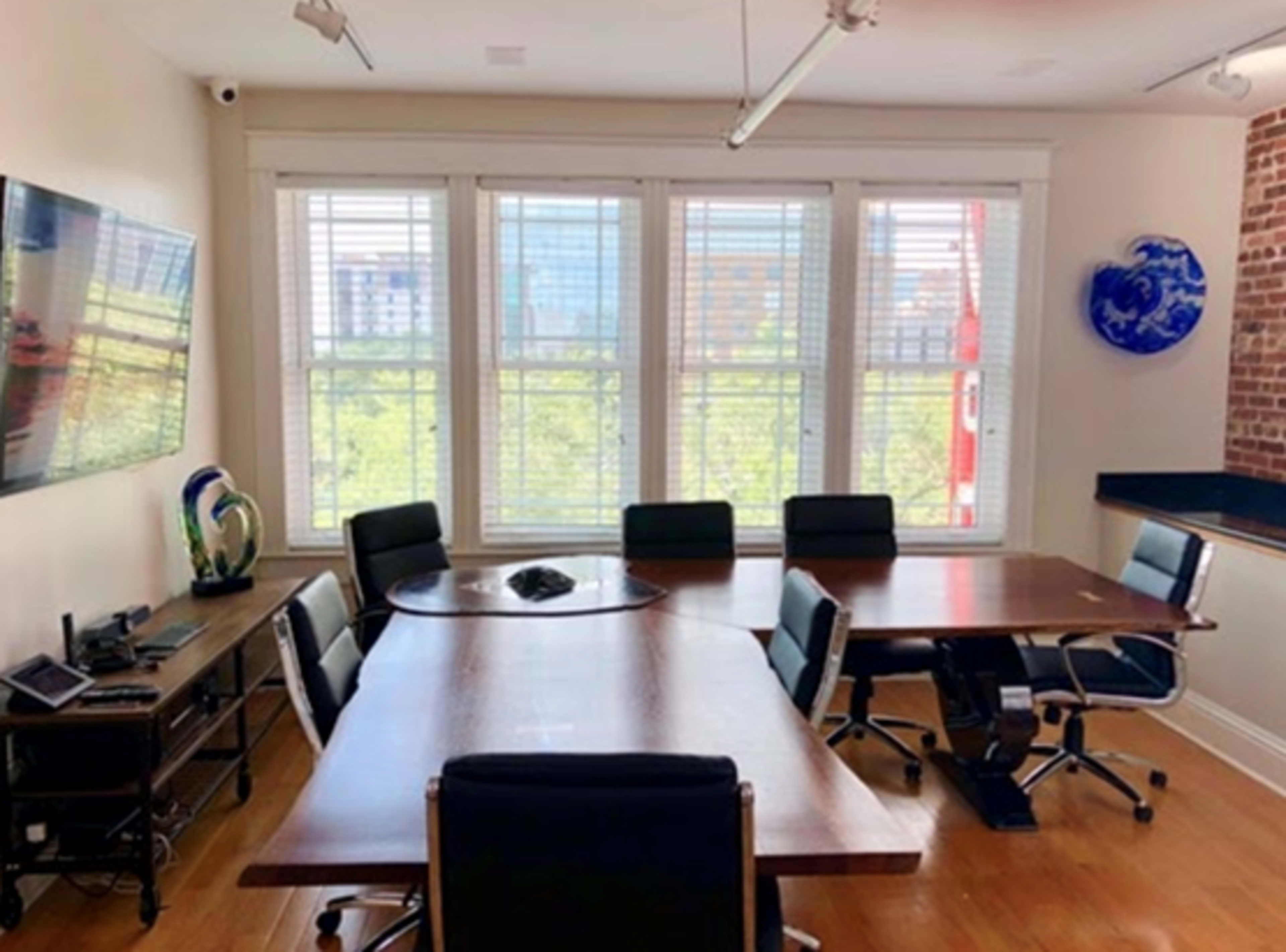A conference room features a large wooden table surrounded by chairs, with a television on the wall and large windows overlooking a greenery-filled view.