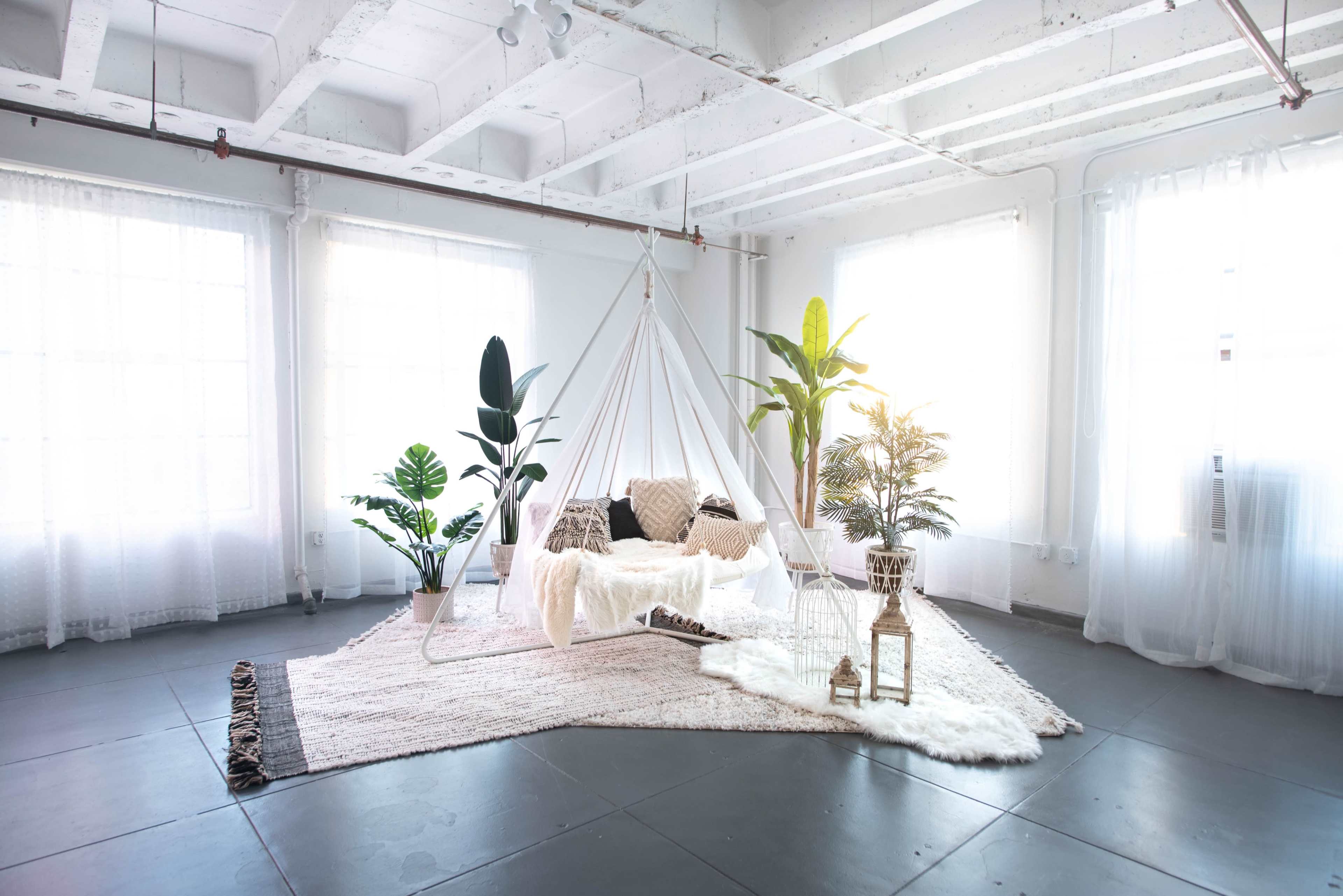 A bright, minimalistic interior space featuring a hanging chair surrounded by various plants on a large, textured rug.