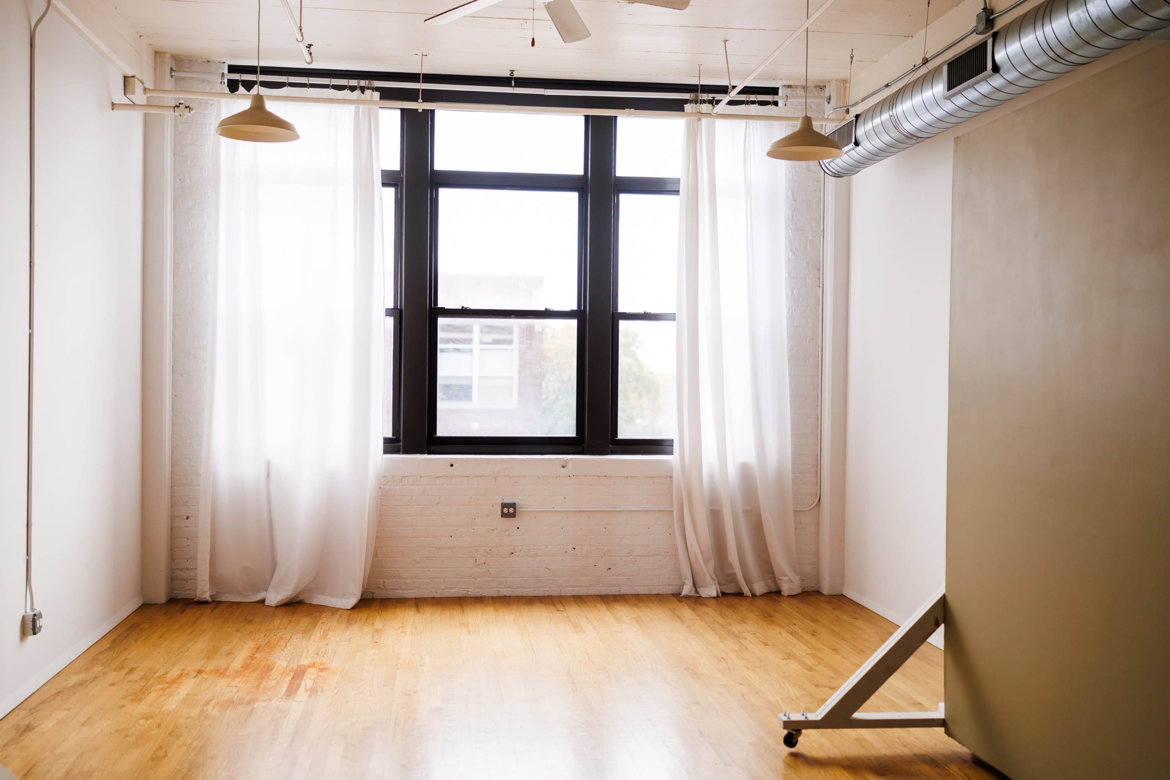 The image shows a minimalist interior space with large windows, sheer curtains, and a hardwood floor.