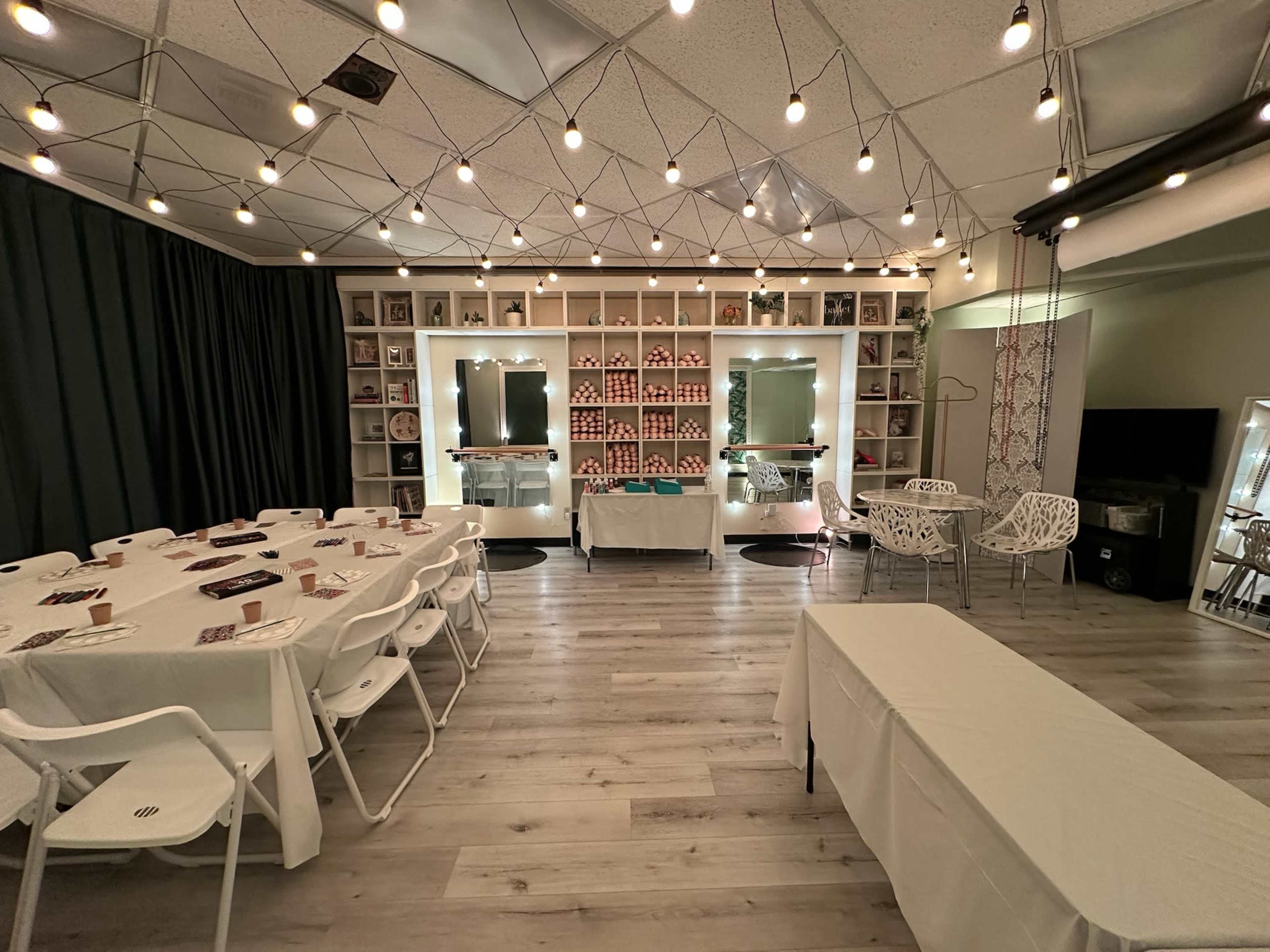 The image shows a well-lit event space with a large table set up for dining, white chairs around it, and a decorative shelving unit featuring various items in the background.