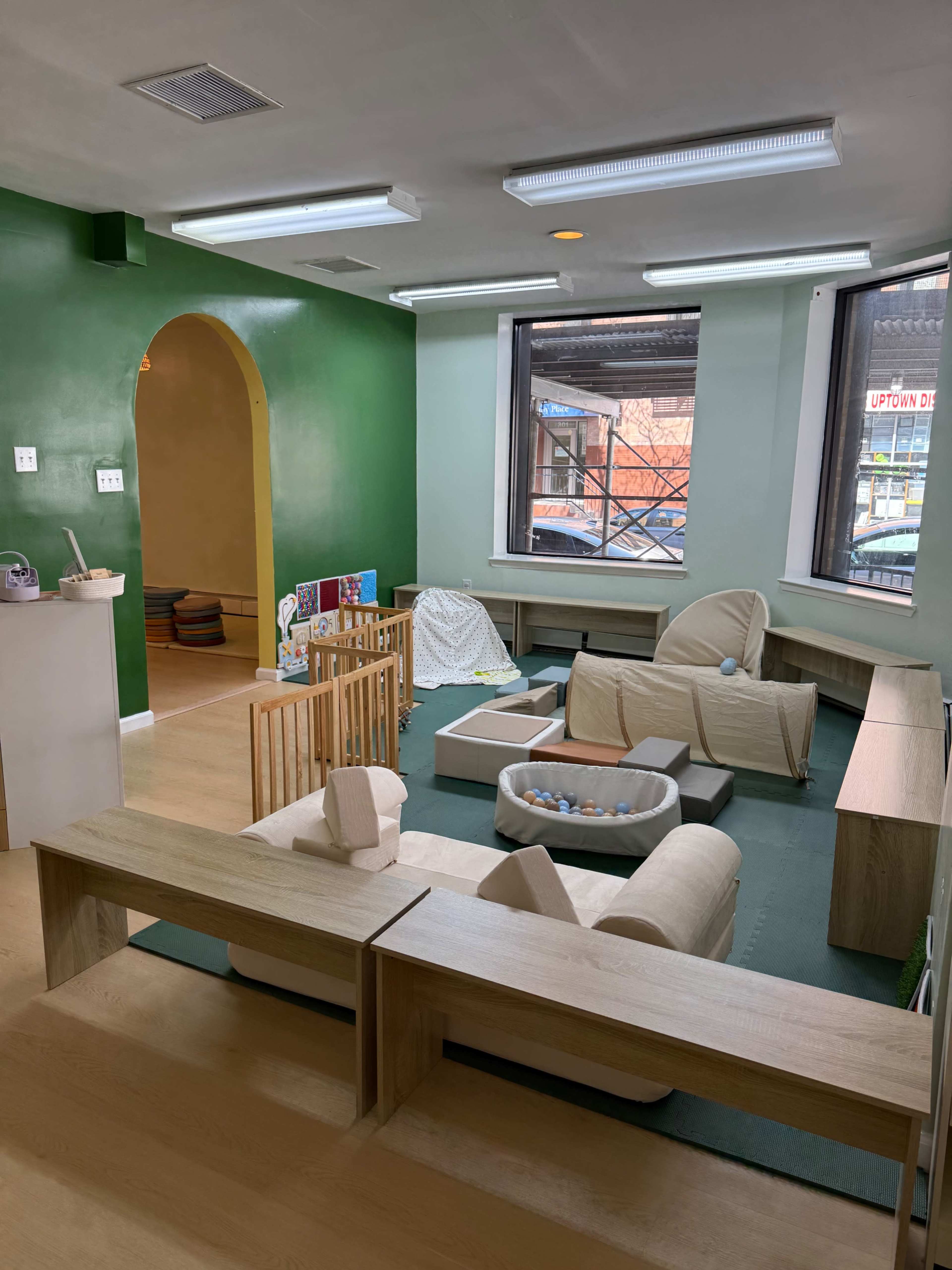 The image shows a brightly colored playroom with soft furniture, a gated area for children to play, and windows allowing natural light to enter.