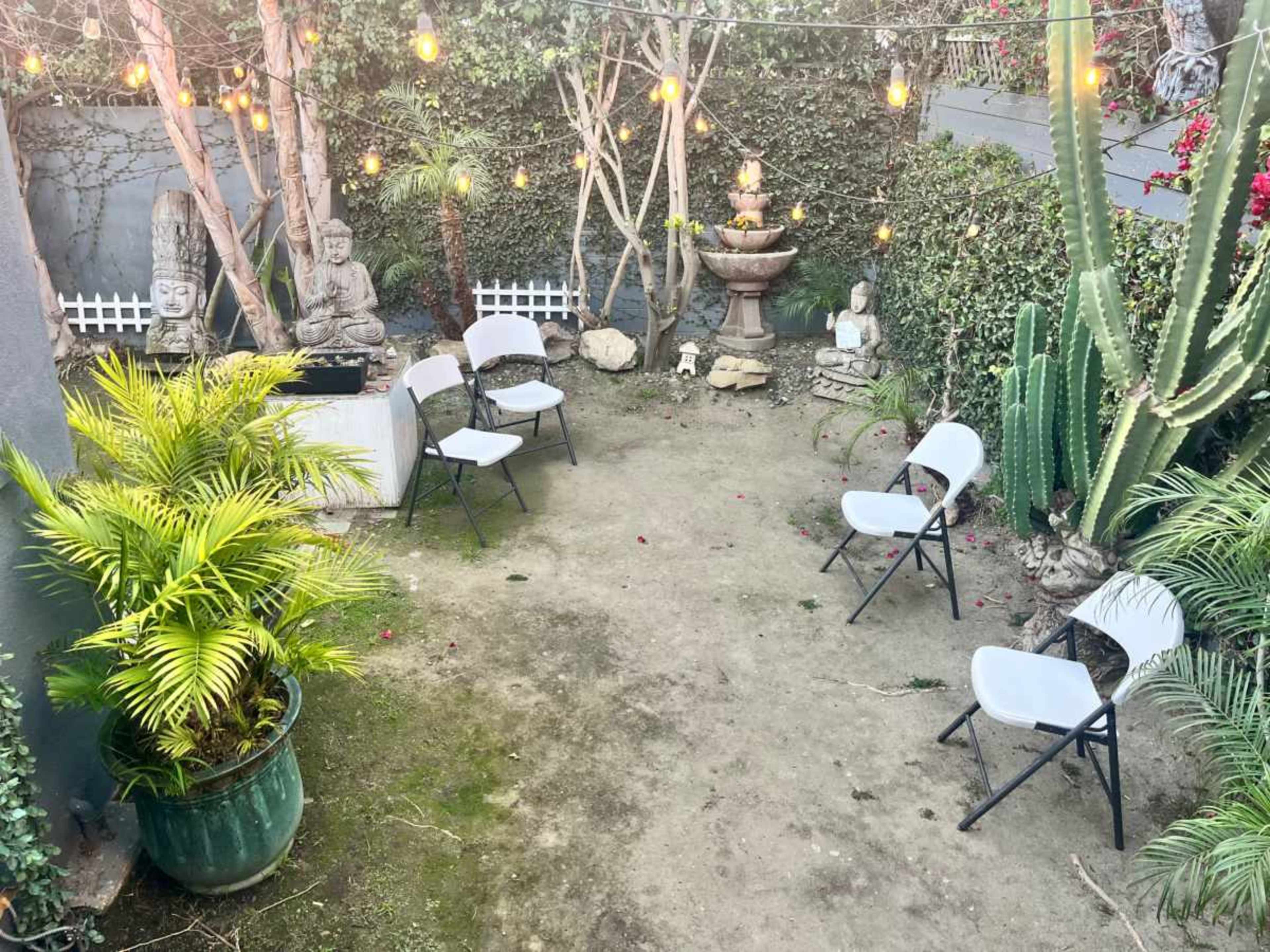 The image shows a small outdoor garden with scattered folding chairs, decorative statues, and string lights overhead amidst greenery and a fountain.