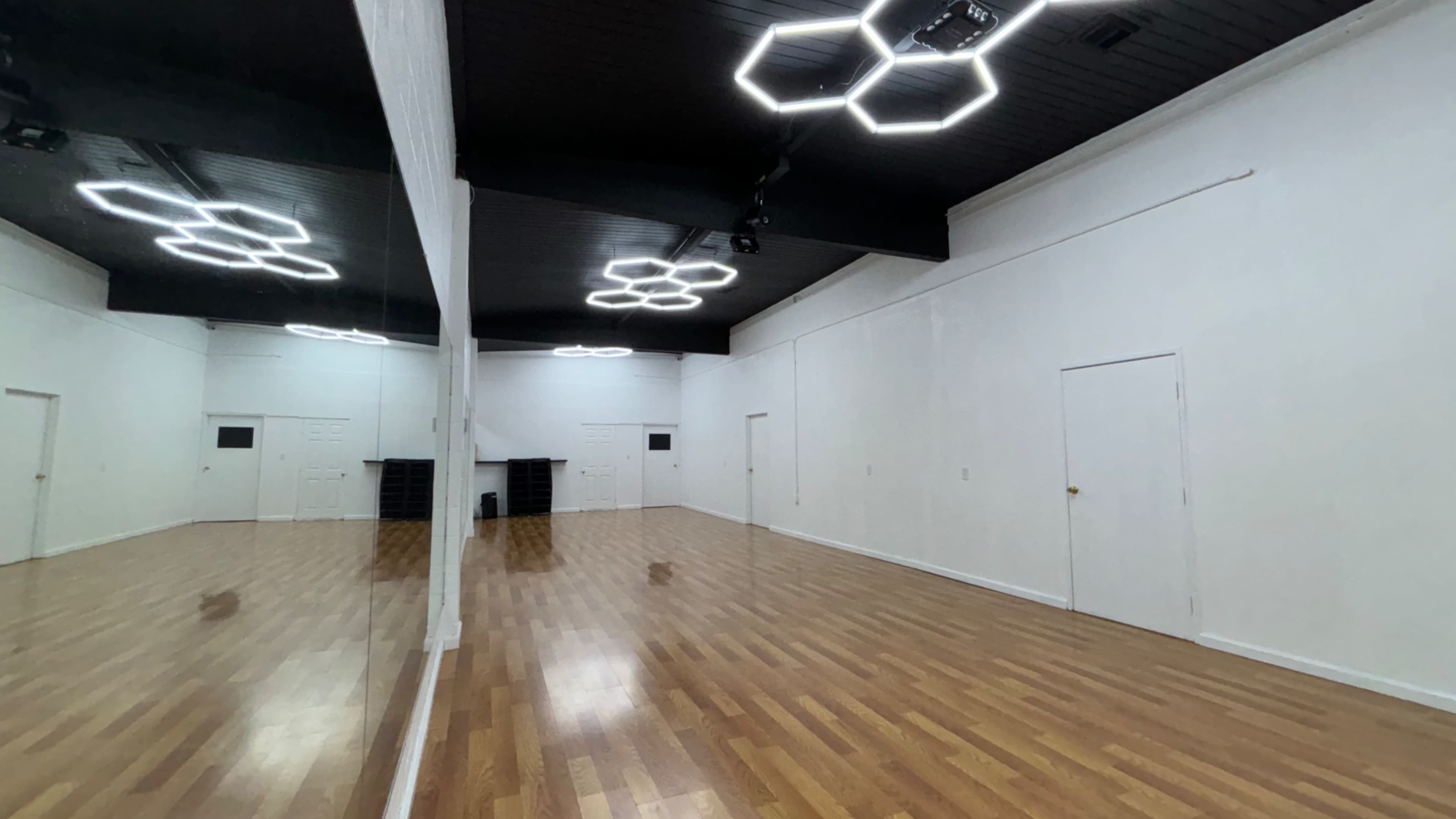 A spacious, empty dance studio with wooden flooring, white walls, and modern light fixtures on the ceiling.