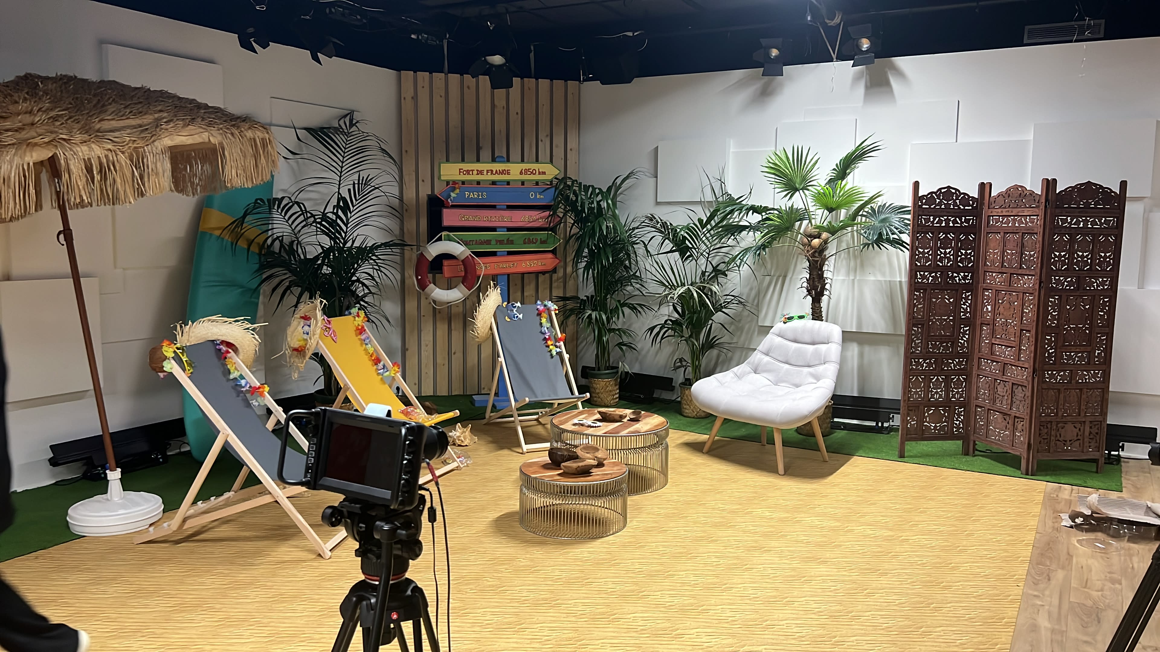 A bright studio setup with colorful lounge chairs, tropical plants, a decorative partition, and a camera on a tripod ready for a shoot.
