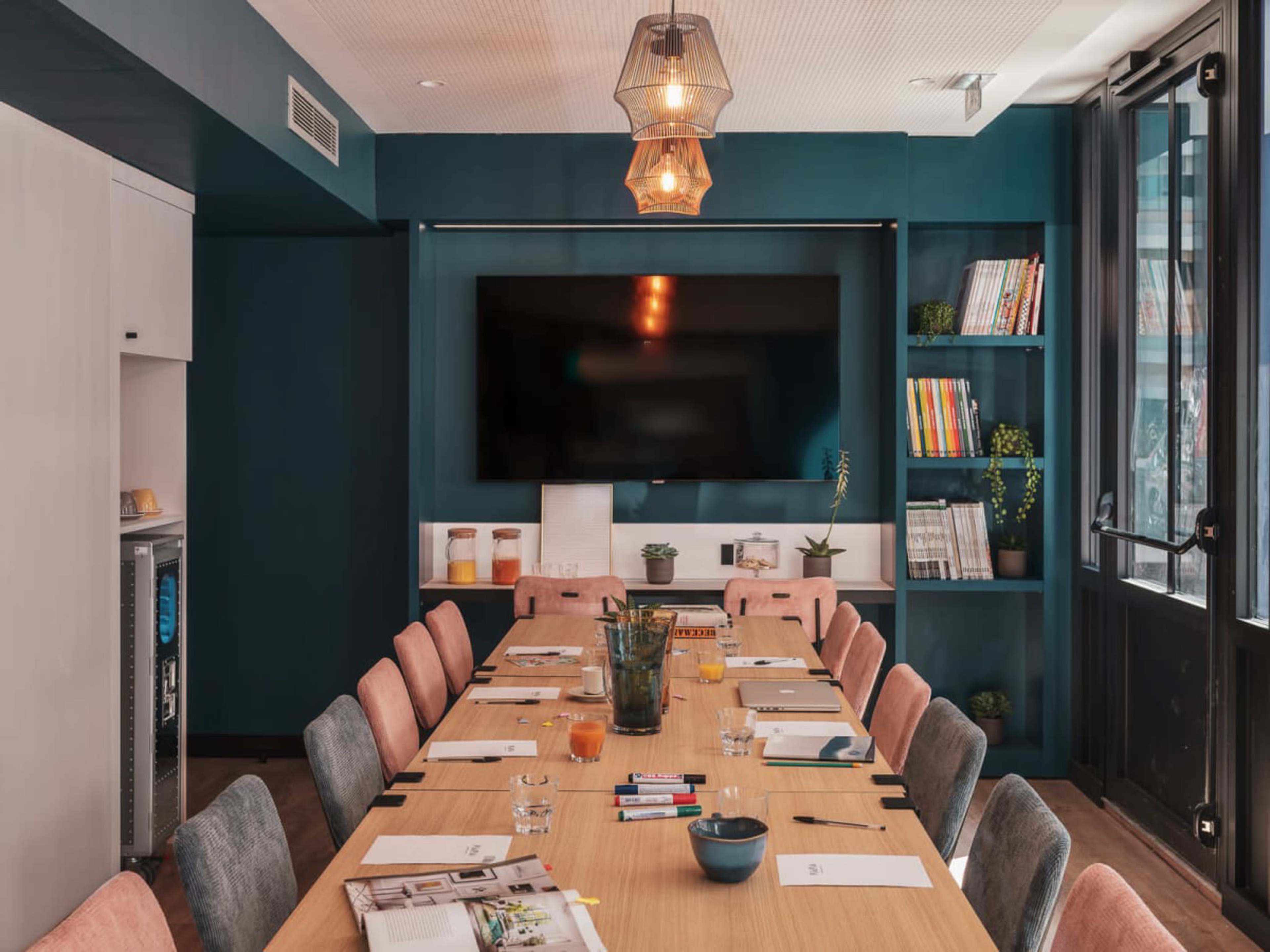 A long wooden conference table with chairs is set up in a room featuring a large television and shelves filled with books and plants.