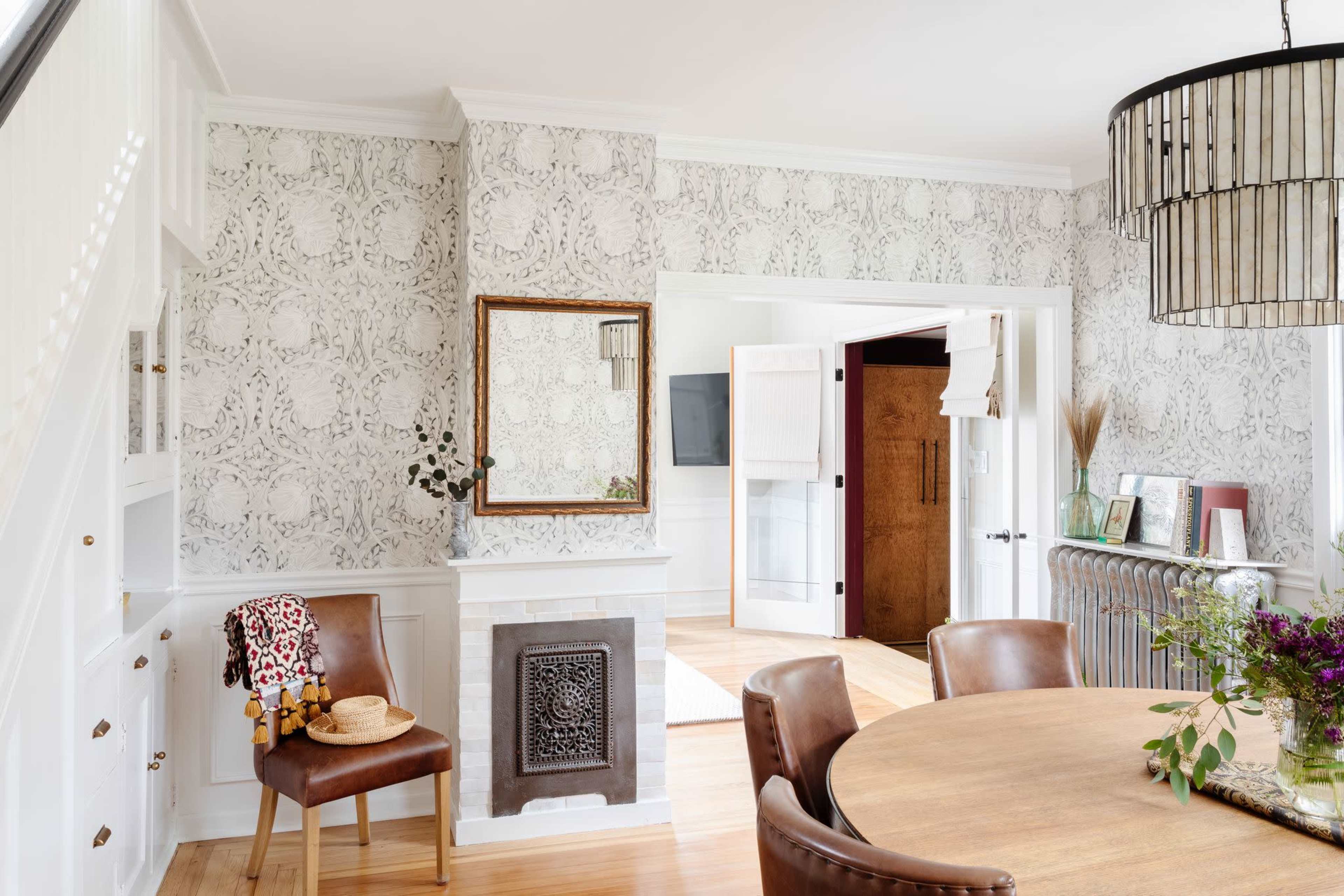 A dining area features a round wooden table surrounded by chairs, with a patterned wallpaper and a decorative mirror on the wall beside a vintage radiator.