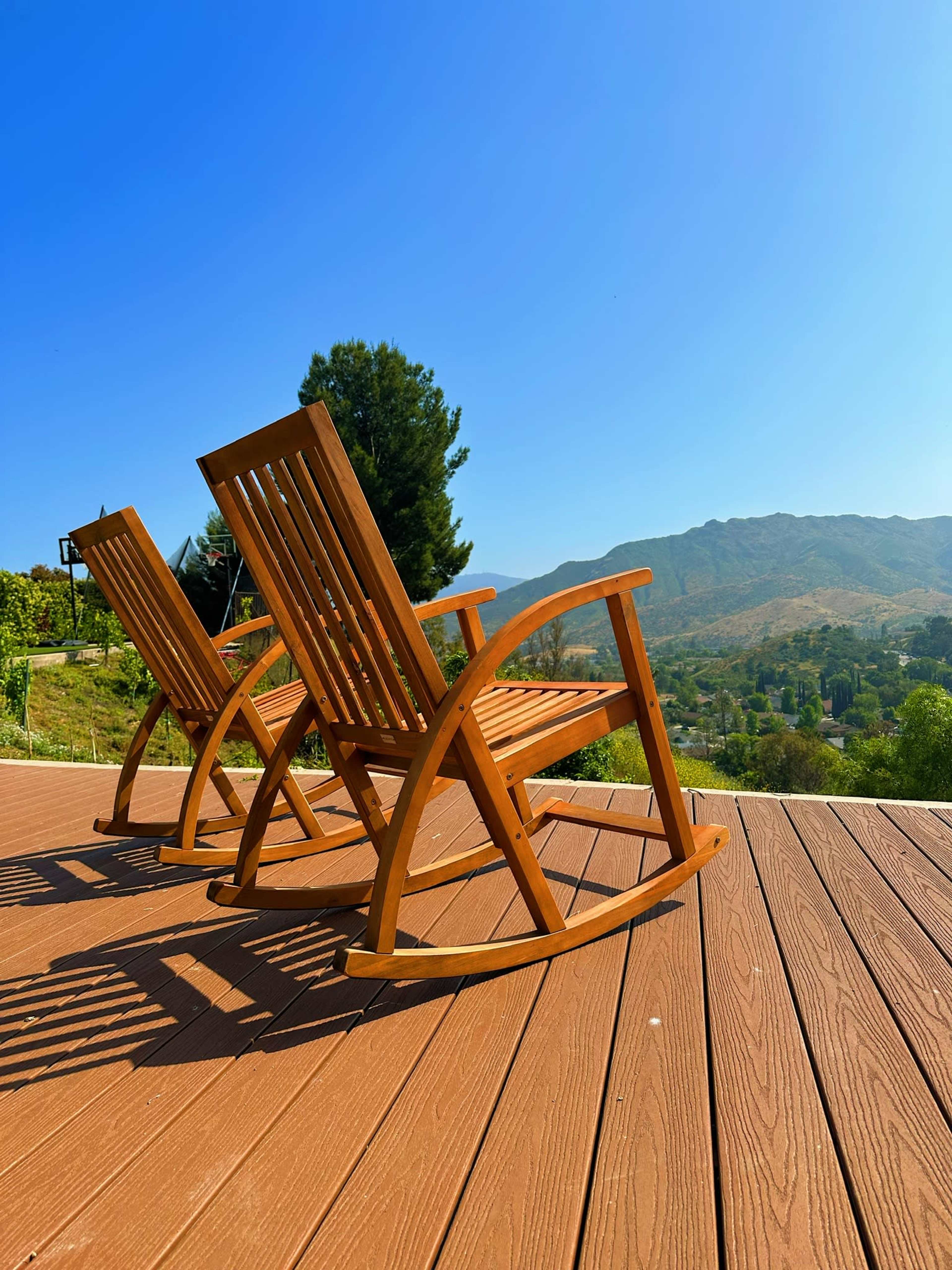 Two wooden rocking chairs are positioned on a patio overlooking a mountainous landscape under a clear blue sky.
