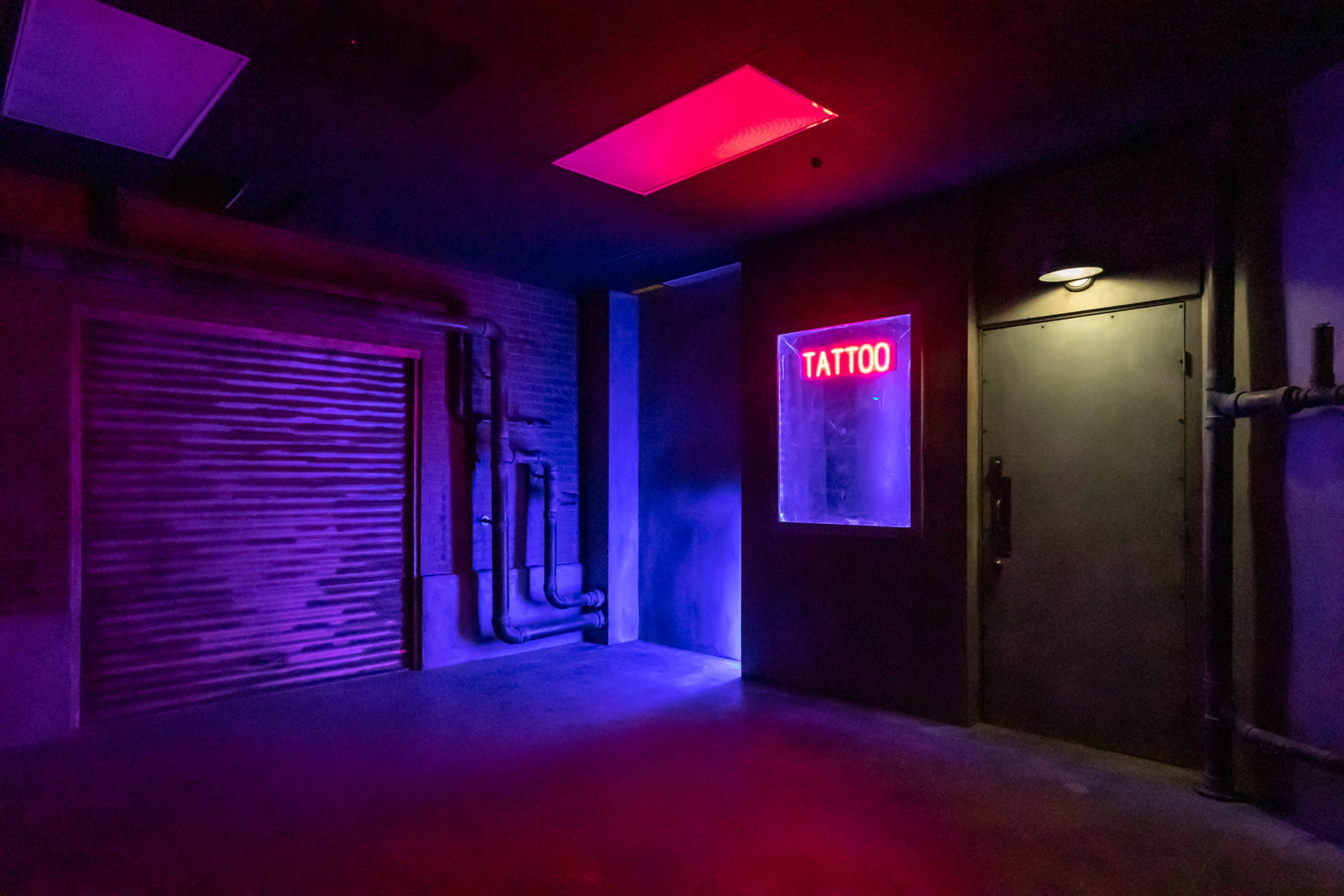 The image shows an underground room illuminated by purple and red lights, featuring a closed door with a "TATTOO" sign and exposed pipes along the wall.