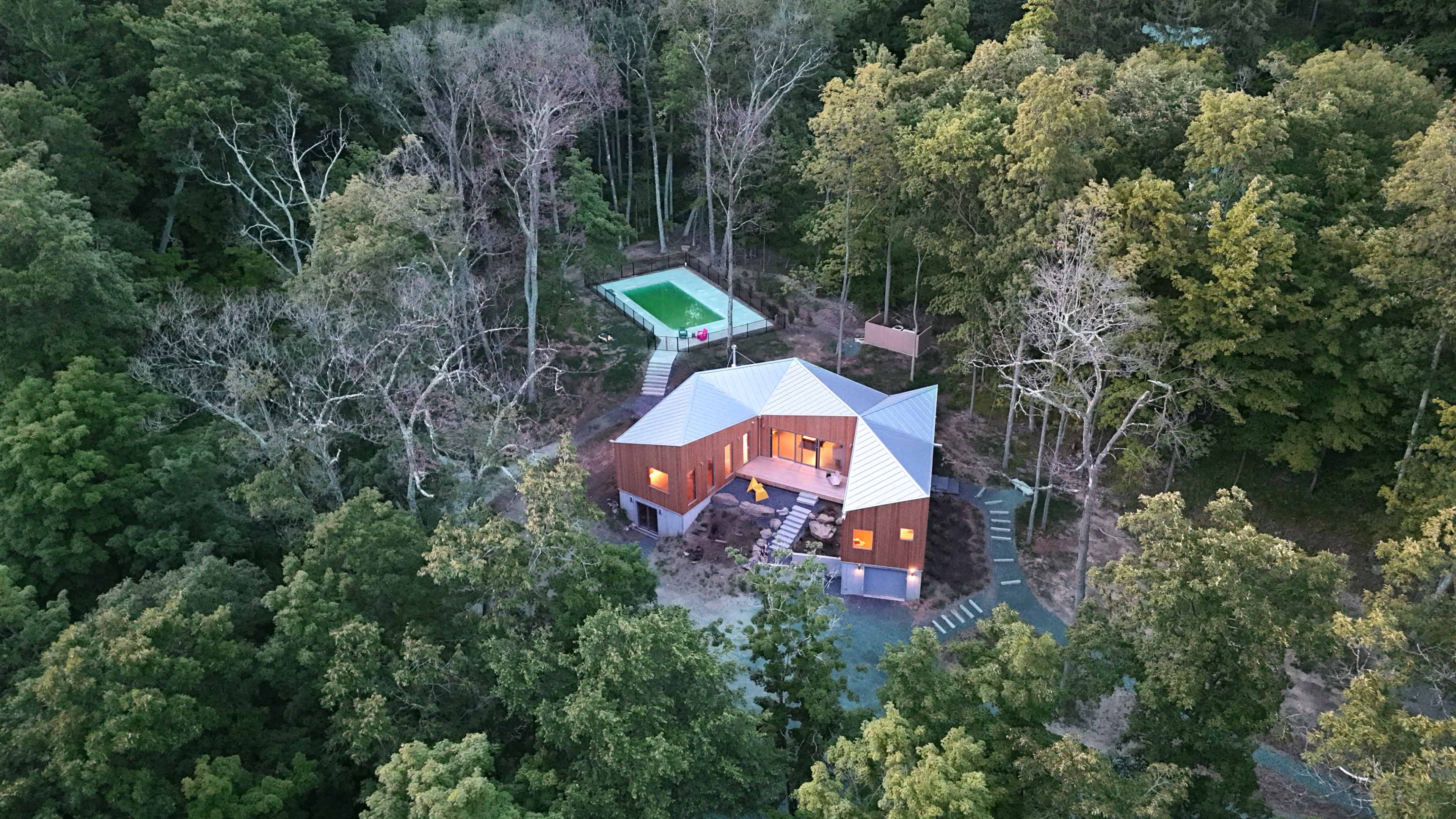 A modern house with a distinctive angular roof is situated amidst a dense forest, featuring a pool nearby.