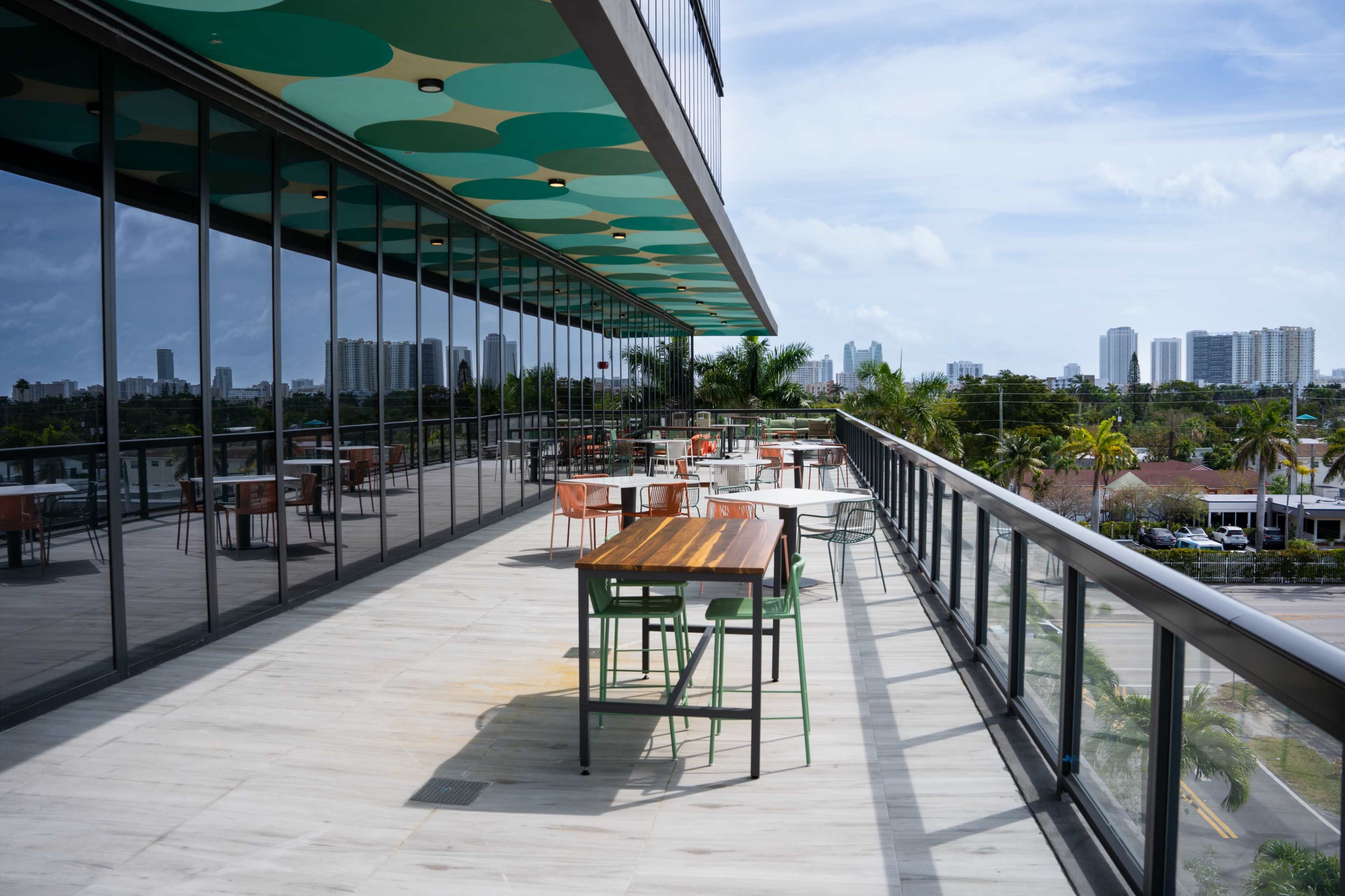 A rooftop terrace features several tables and chairs with a view of city buildings and a clear blue sky.