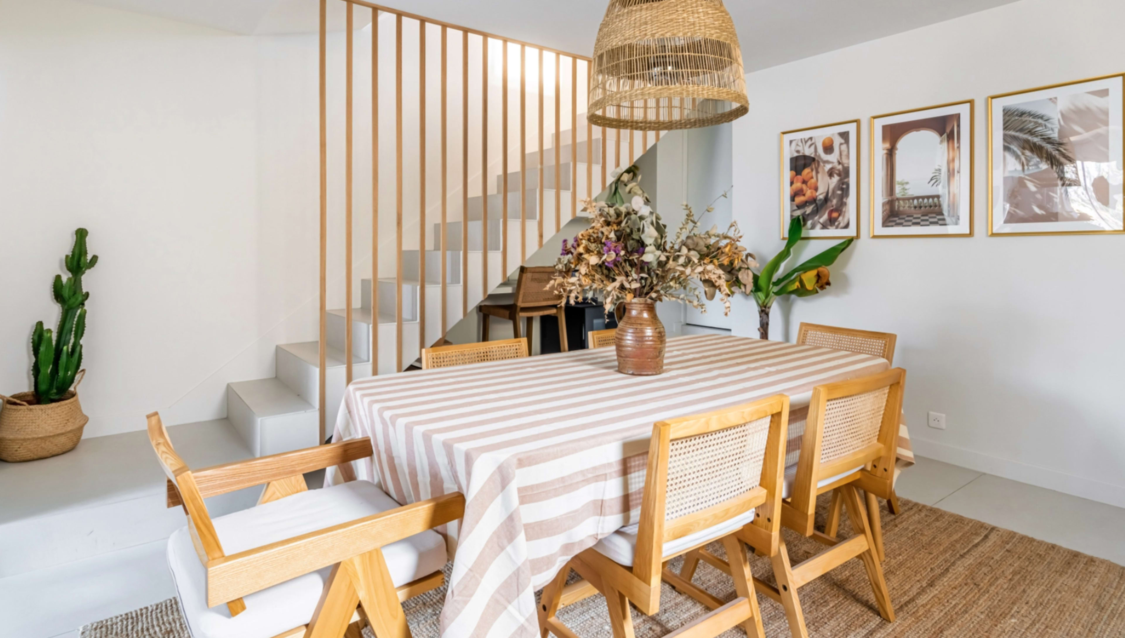 A dining area features a striped tablecloth on a wooden table surrounded by six chairs, with a staircase and decorative elements in the background.
