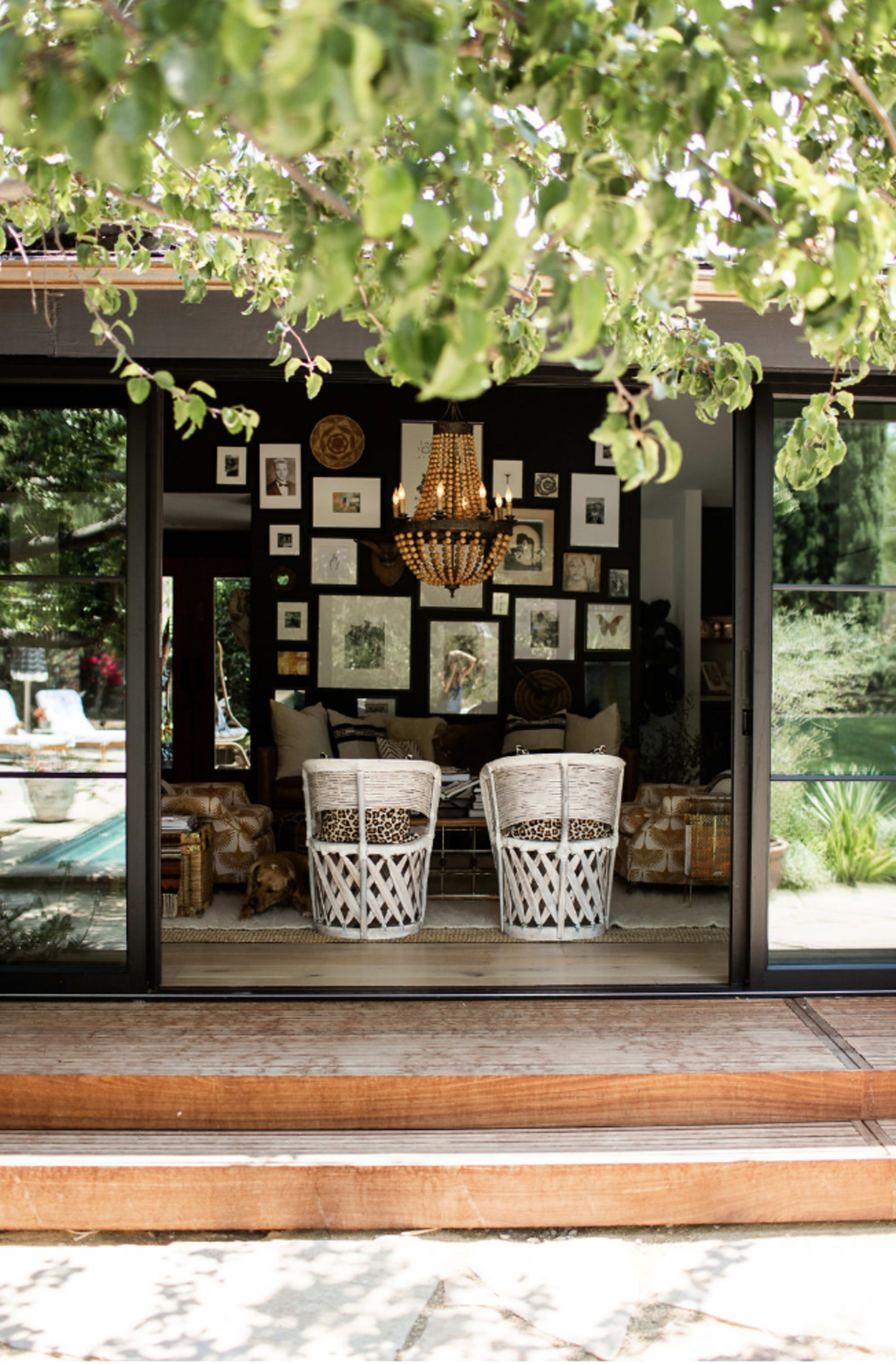 A cozy living space is visible through a large glass door, featuring two white wicker chairs, a chandelier, and walls adorned with framed artwork.