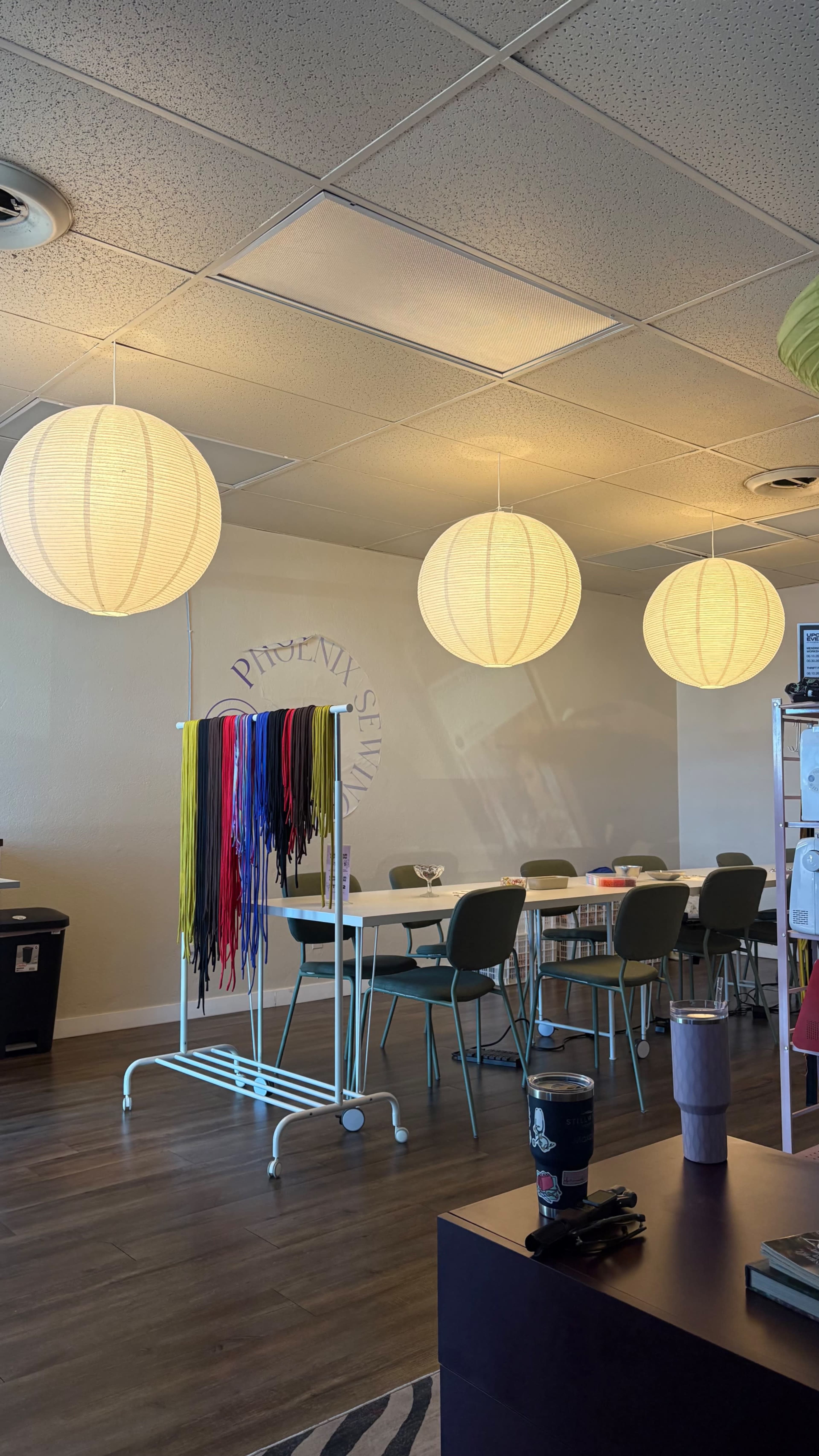 The image shows a well-lit room with several round paper lanterns hanging overhead, a table and chairs arranged for gatherings, and a colorful garment rack in the foreground.