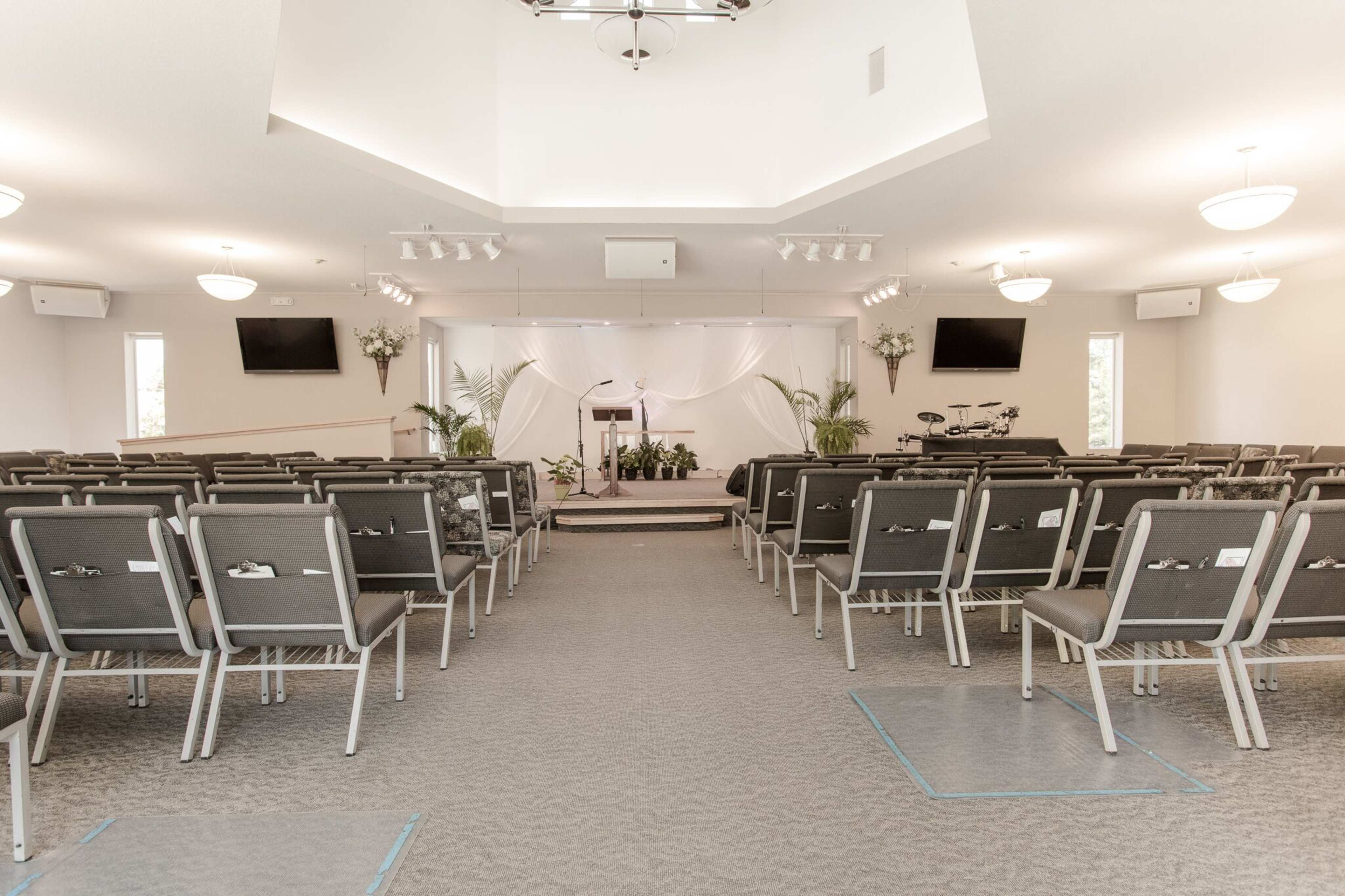 A spacious interior of a church with rows of gray chairs facing a podium surrounded by plants and decorative white drapery.