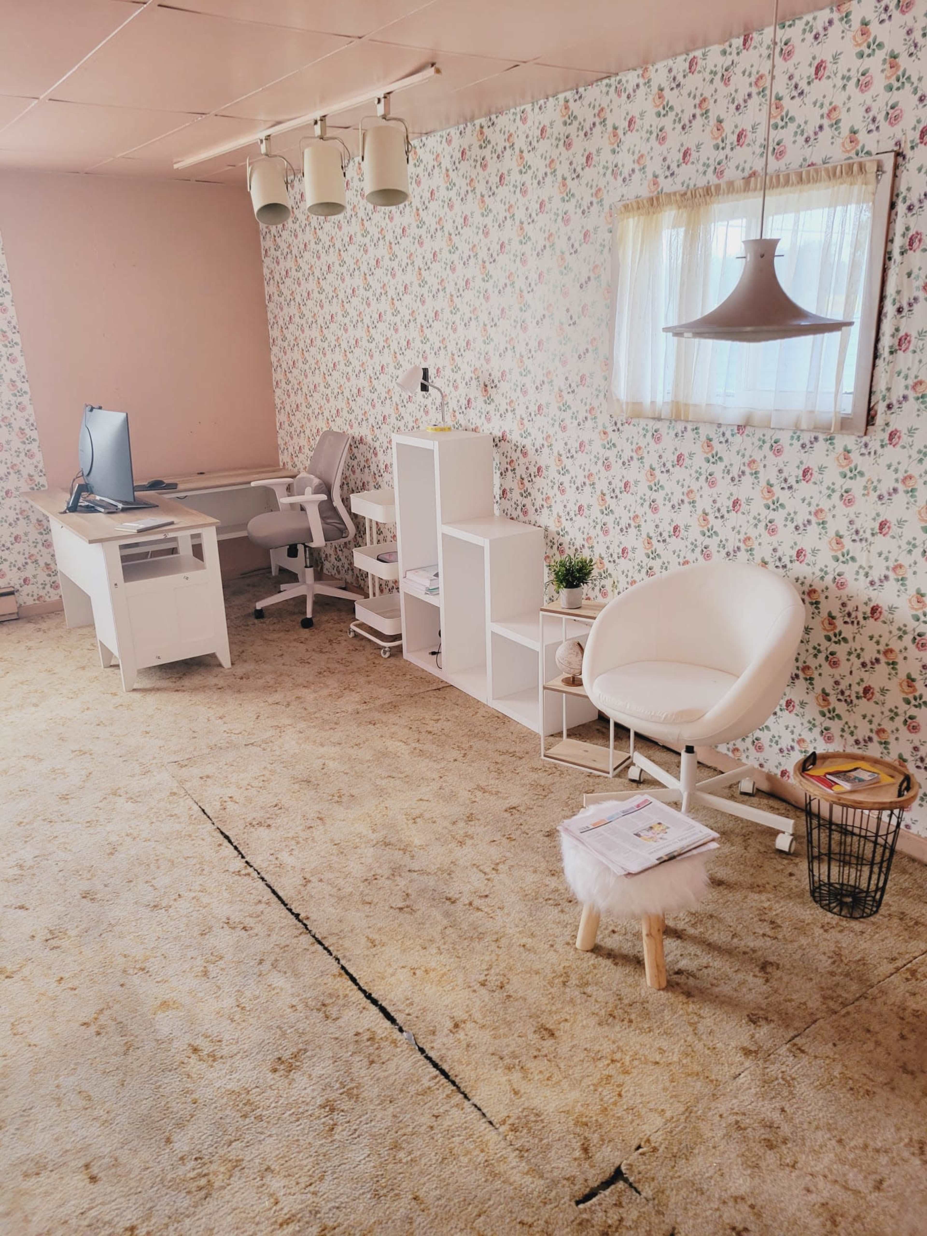 A room with floral wallpaper features a desk with a computer, a white chair, a small table, and storage shelves, all arranged on a bare carpet.