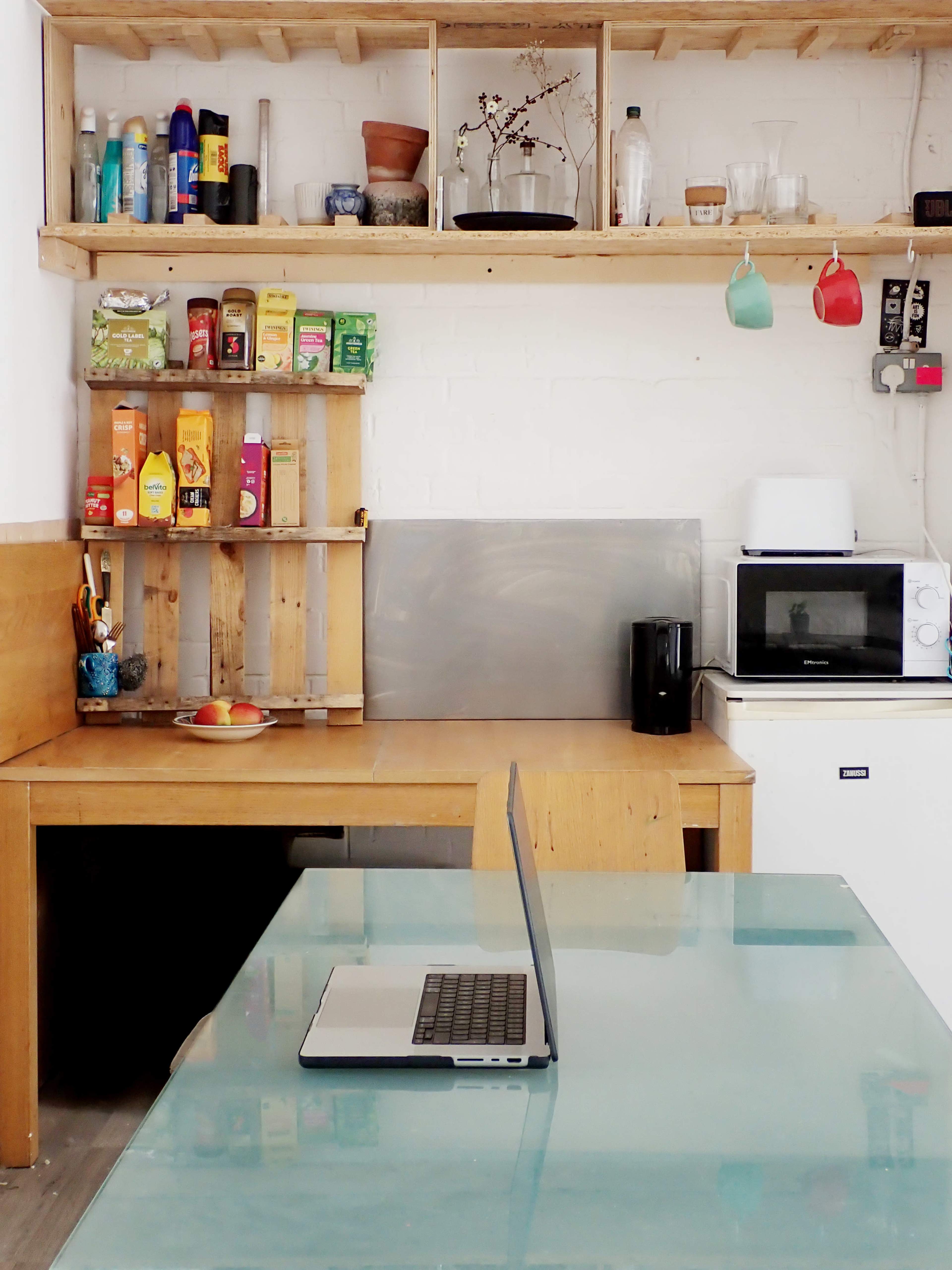 The image shows a small kitchen with a wooden table, a laptop on the table, shelves filled with various food items and kitchen supplies, and a microwave and refrigerator in the corner.