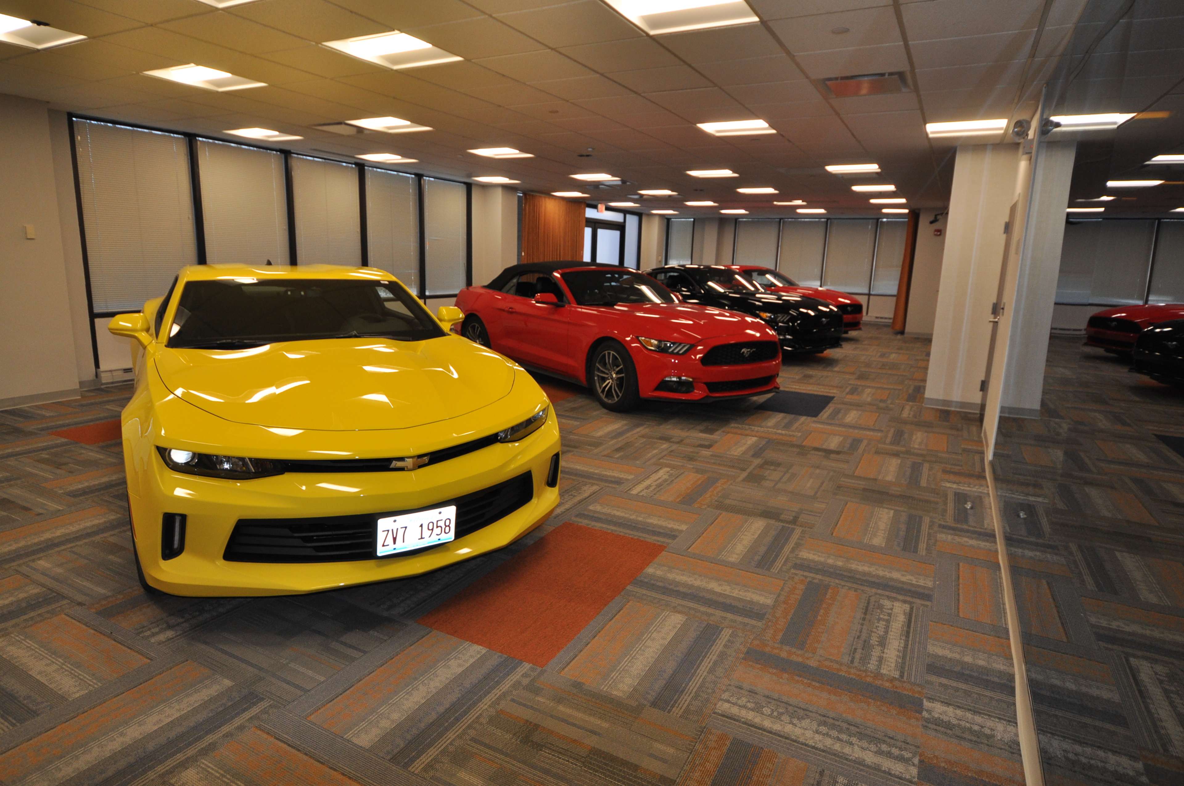The image shows a yellow sports car and a red car parked in a well-lit office space with polished floors and large windows.