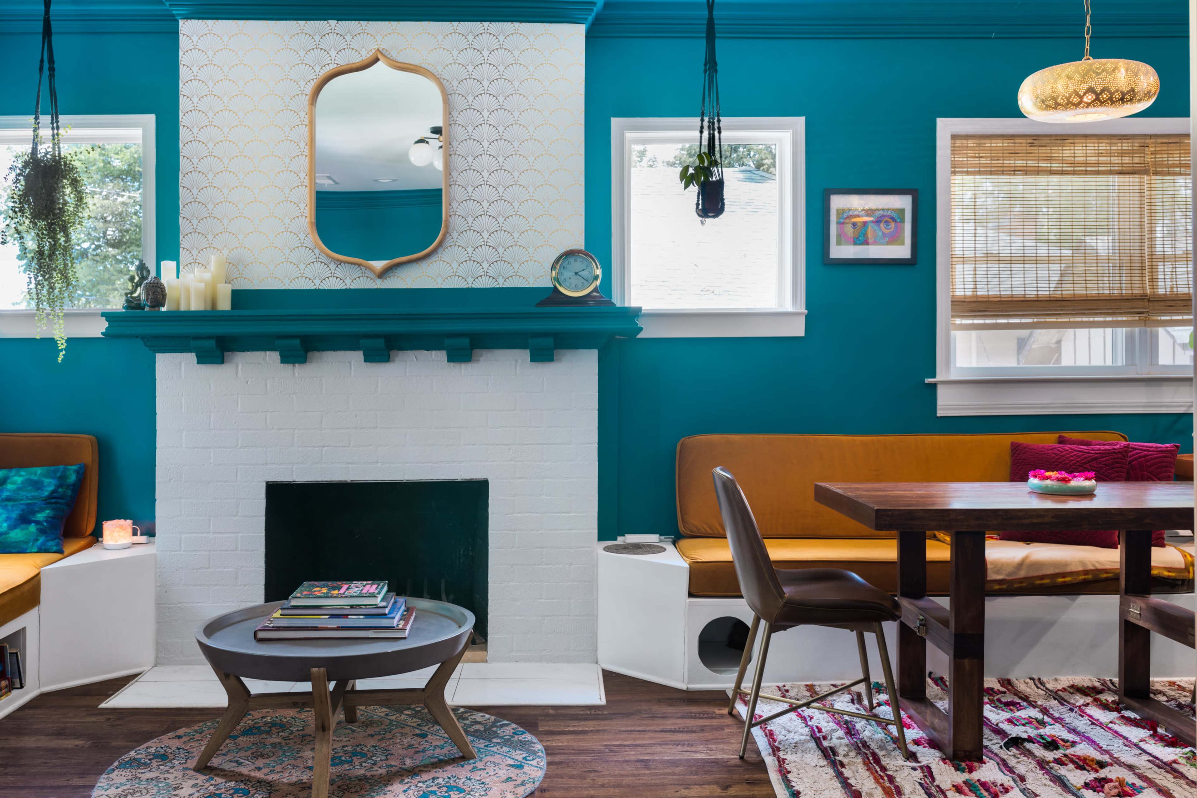 The image shows a bright teal living room featuring a fireplace, a wooden table, seating areas, and large windows with shades.