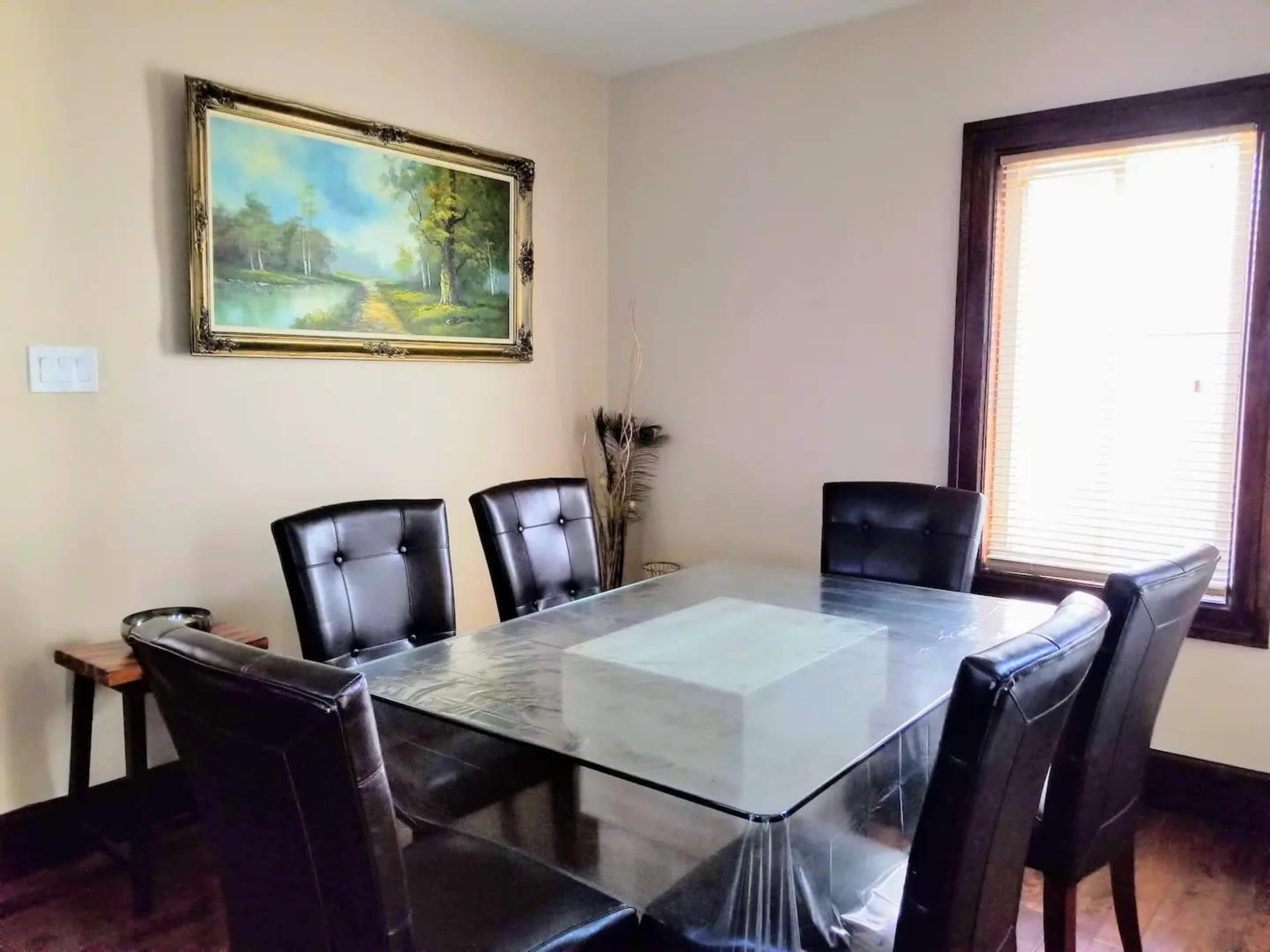 A dining room features a glass table surrounded by six black leather chairs, with a painting of a landscape on the wall and a window letting in natural light.