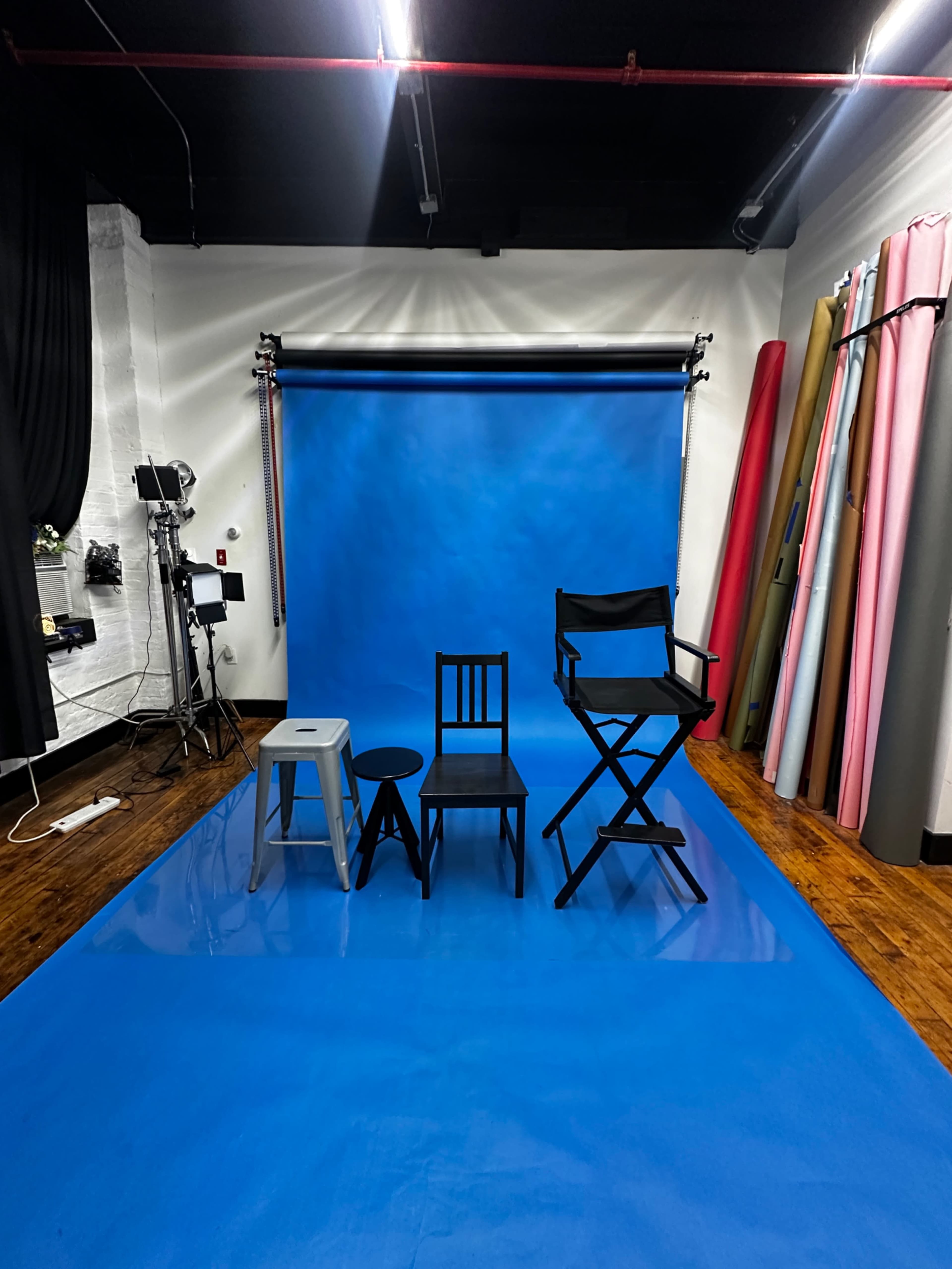 The image shows a photography studio with a blue backdrop, four various chairs positioned on a blue floor, and colorful rolls of backdrop paper along the walls.