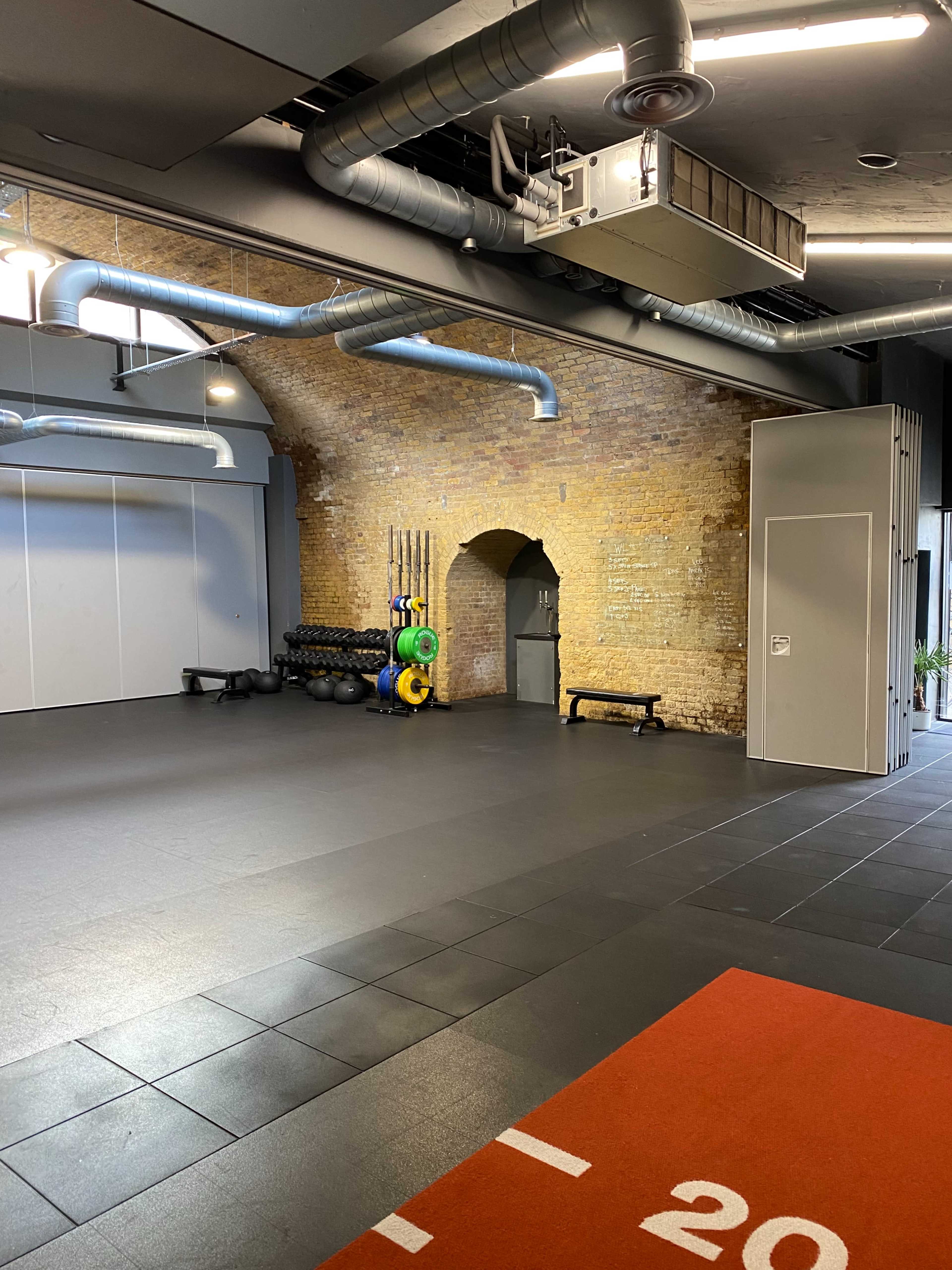An empty gym interior with brick walls, fitness equipment stacked in one corner, and gray flooring.