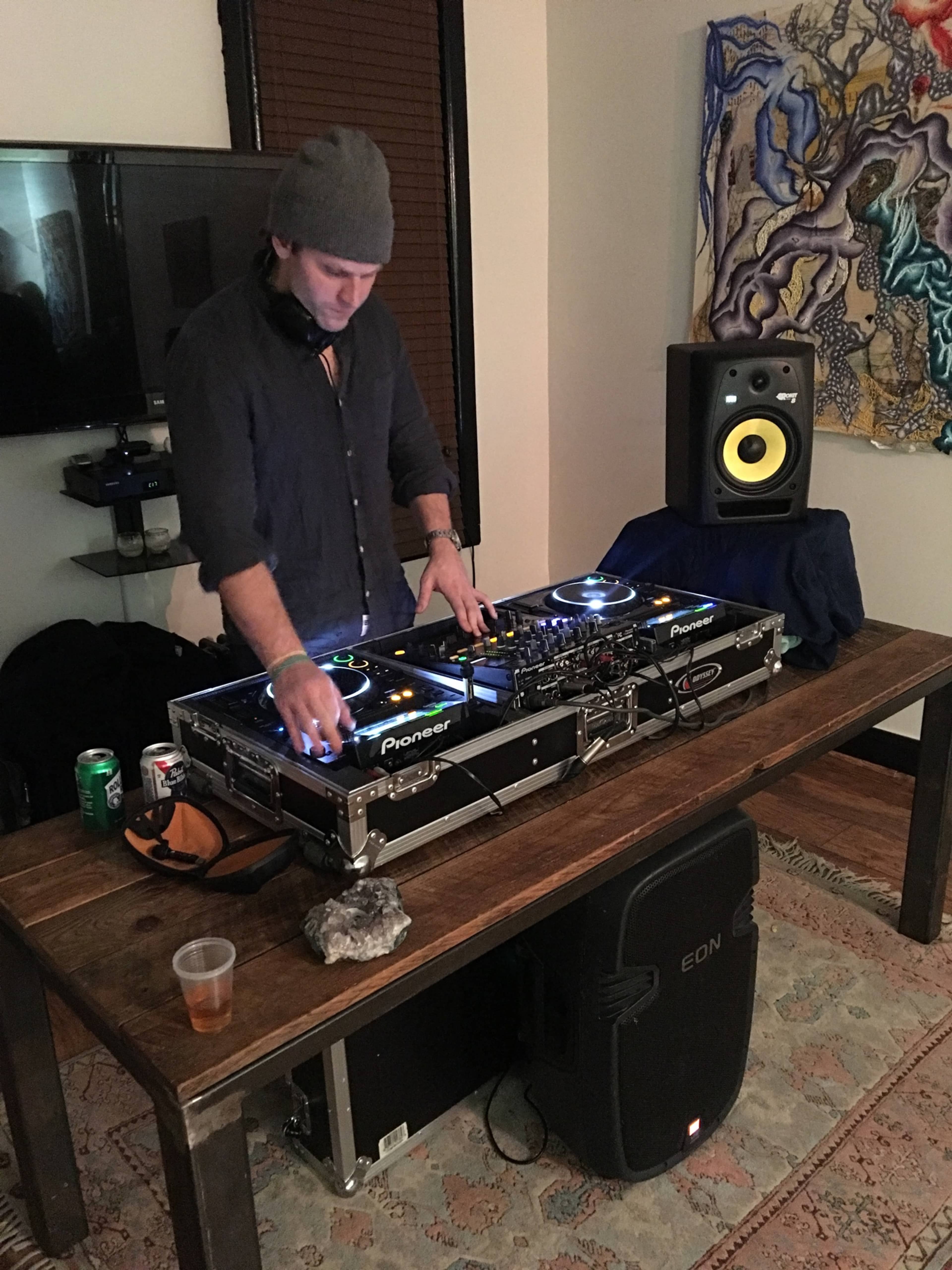 A DJ is standing behind a table with turntables and mixing equipment while a speaker and some drinks are positioned nearby.
