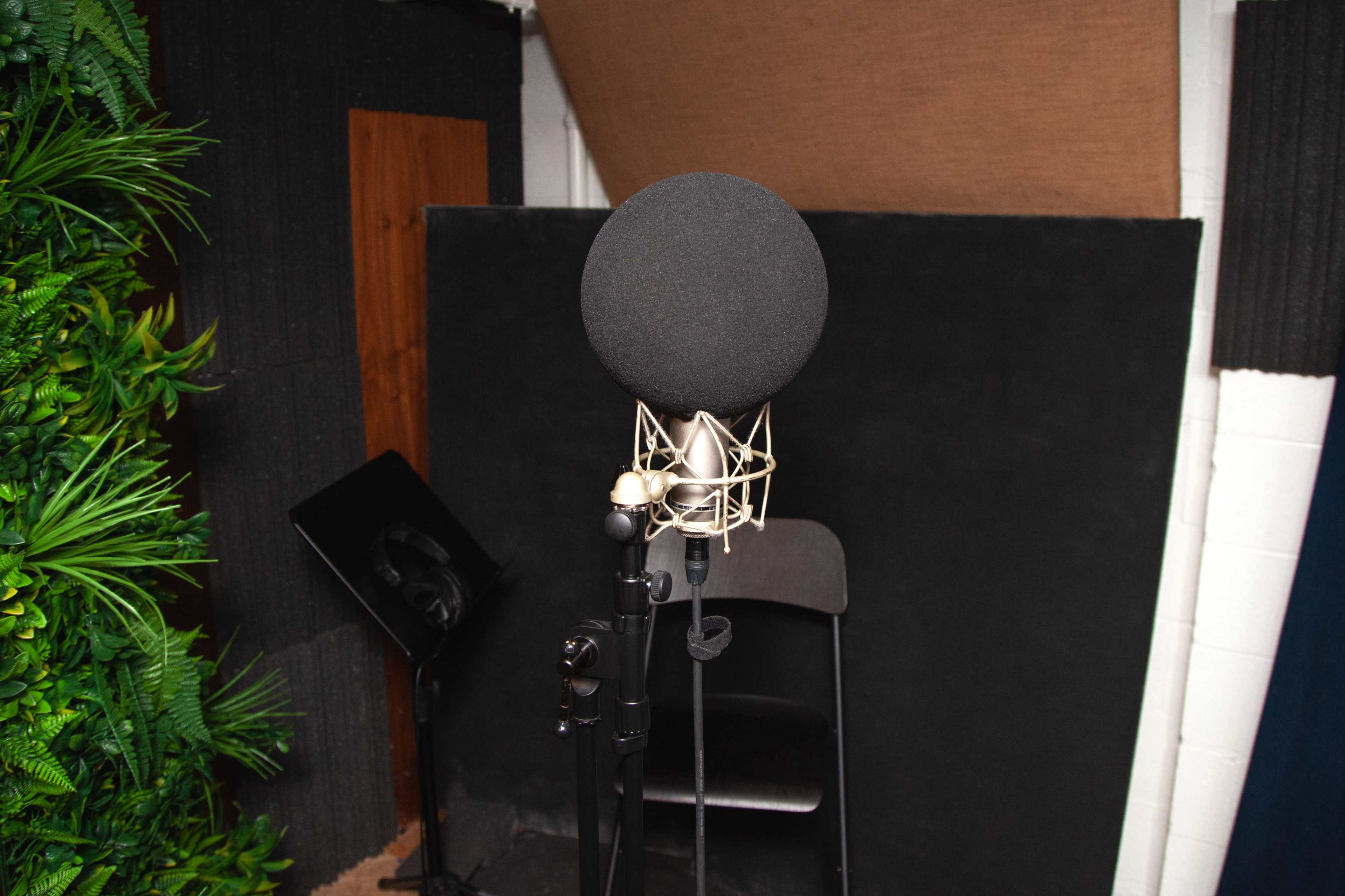 A black microphone stands on a tripod in a soundproof recording studio with dark walls and a chair nearby.