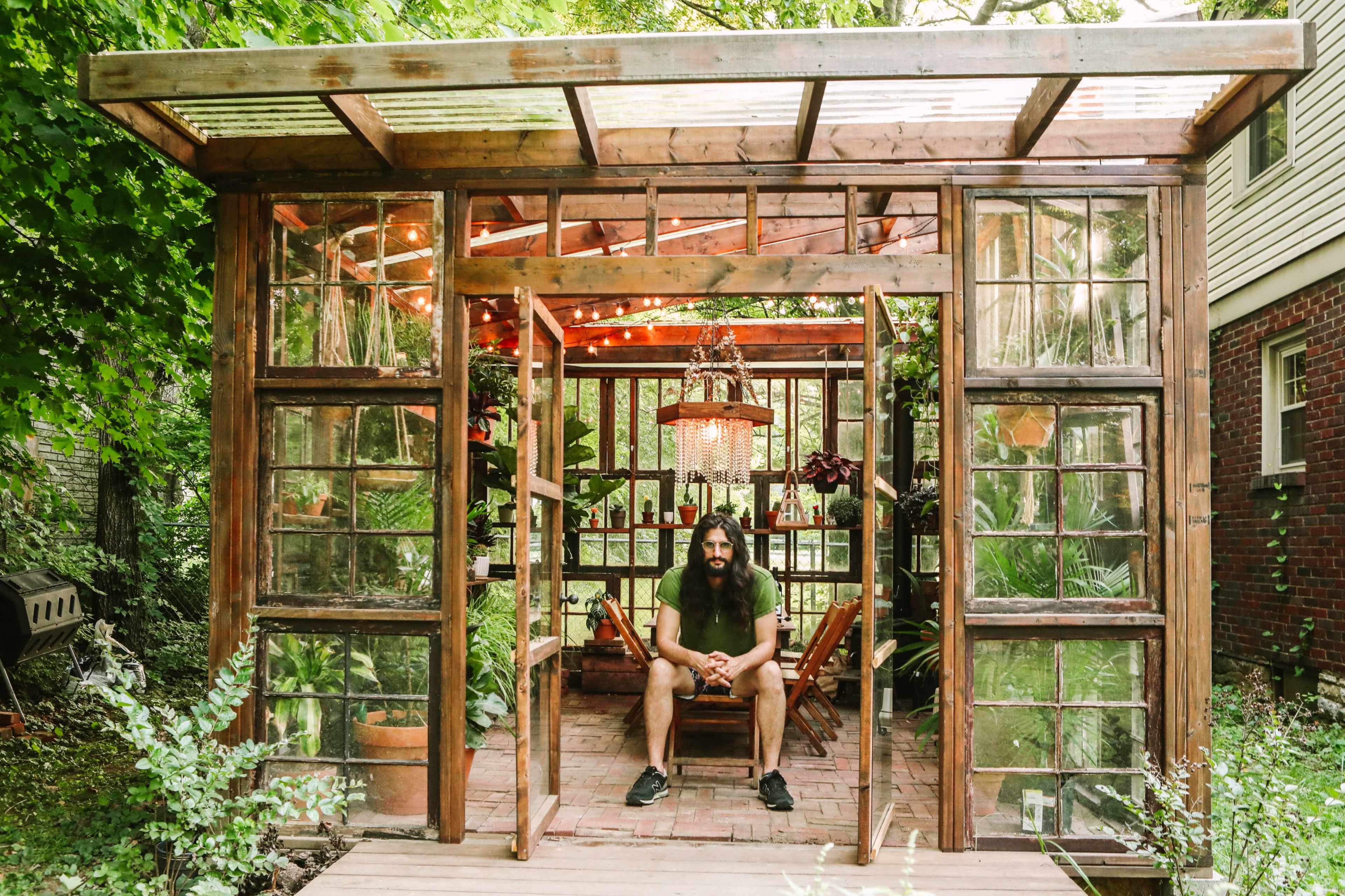 A man with long hair sits inside a wooden greenhouse filled with plants and decorated with string lights.