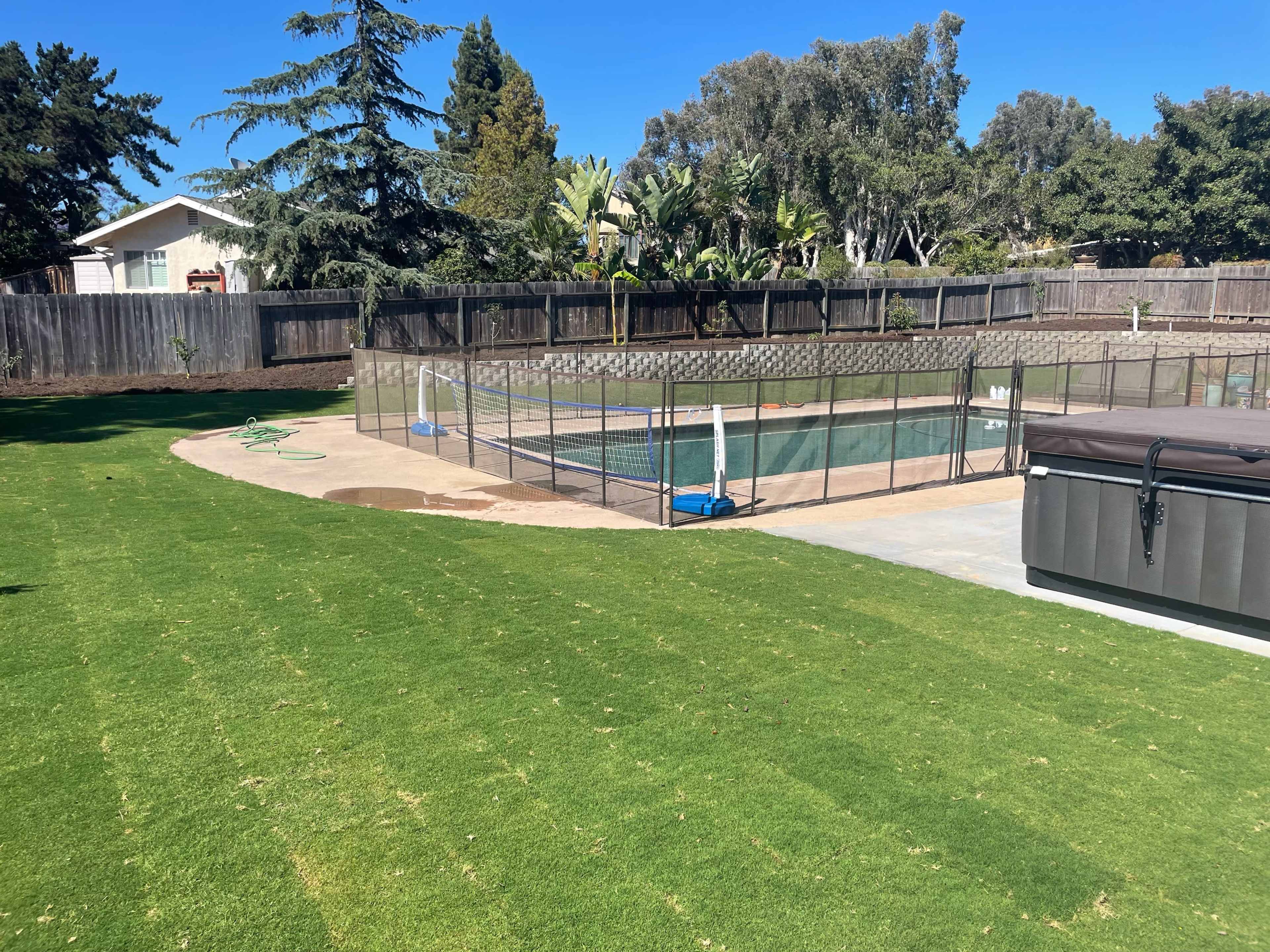 The image shows a backyard with a green lawn, a fenced swimming pool area, and a hot tub.