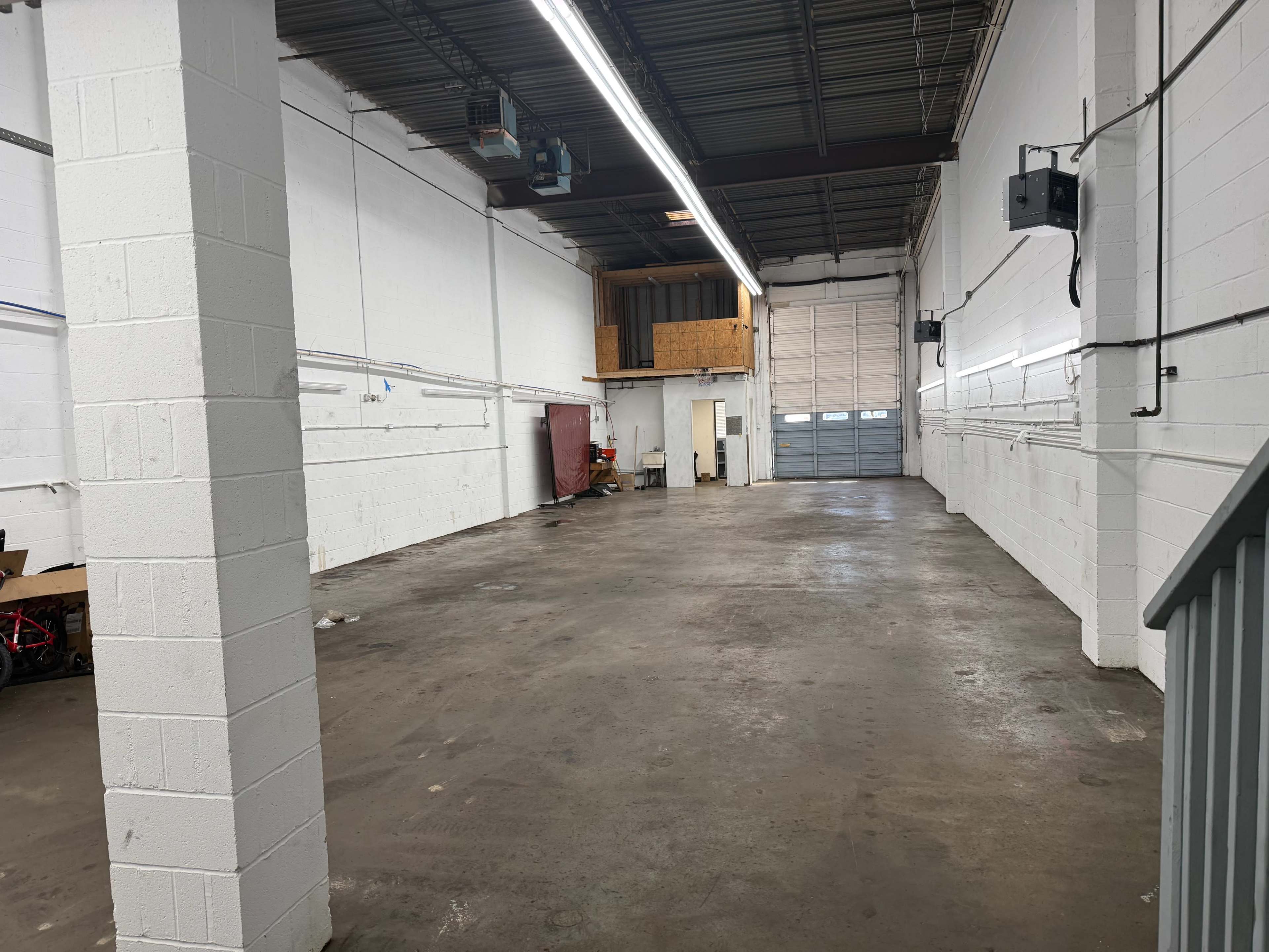 The image shows an empty industrial space with concrete flooring, white cinder block walls, and a large garage door at one end.