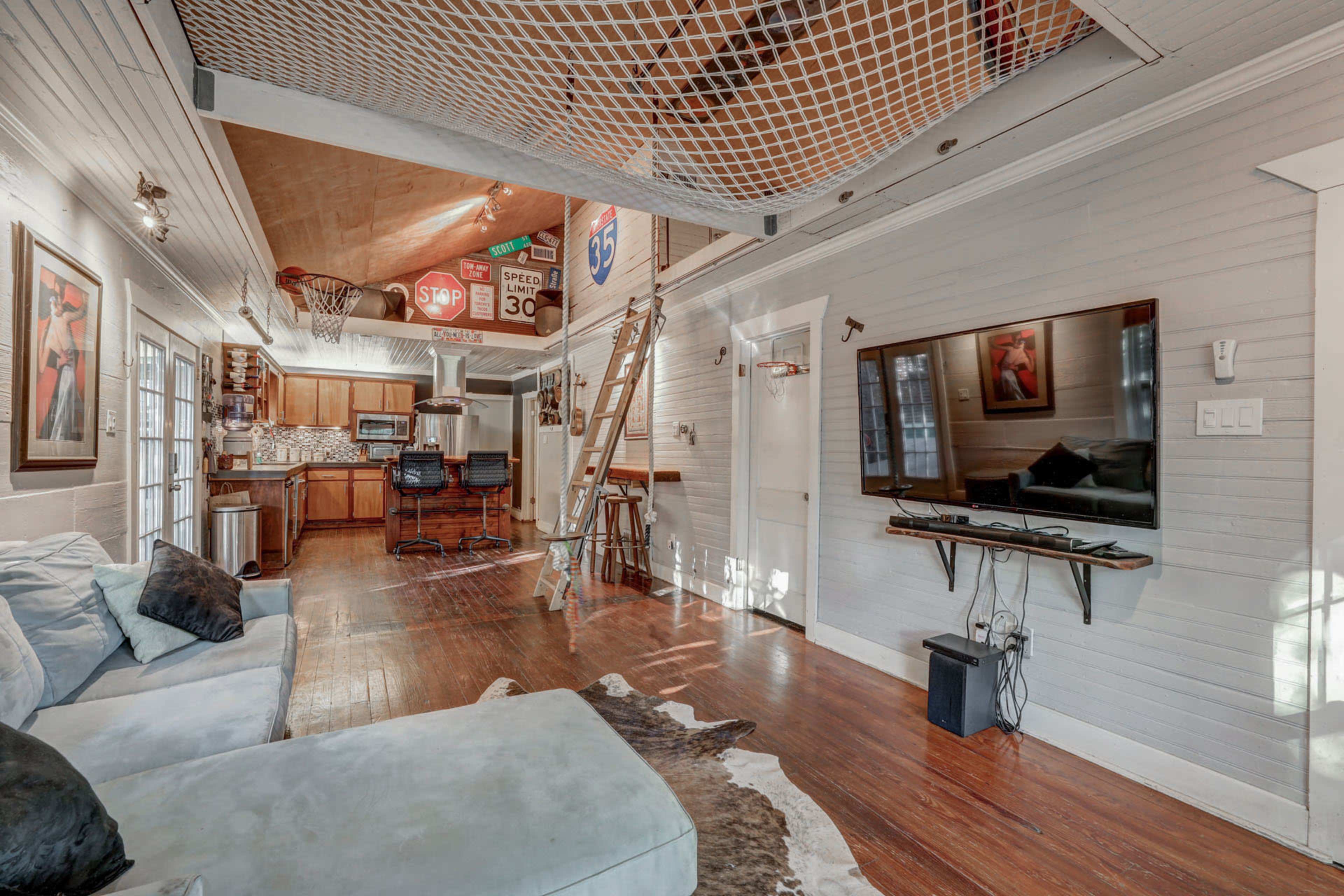 A spacious living area featuring a light-colored couch, a ladder leading to a lofted space, and various signs hanging from the ceiling, with a kitchen visible in the background.